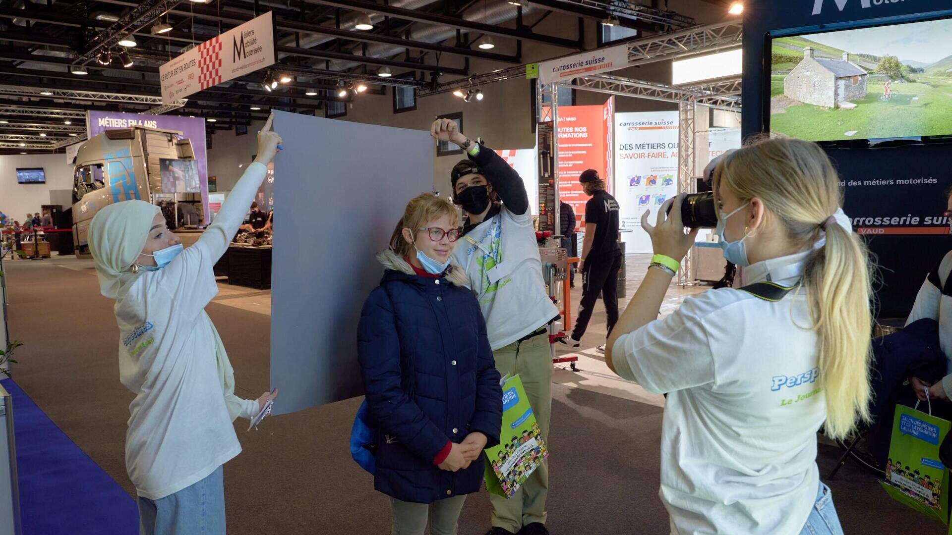 Trois jeunes gens photographient une femme lors d'un événement en intérieur. L'un tient une toile de fond grise, l'autre un appareil photo, et tous portent des masques et des chemises blanches. Des stands d'exposition et des bannières sont visibles à l'arrière-plan.