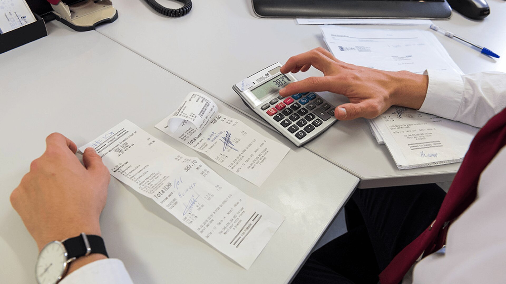 Une personne en tenue de travail utilise une calculatrice et tient des reçus, travaillant à un bureau avec des documents, un stylo bleu et un téléphone.