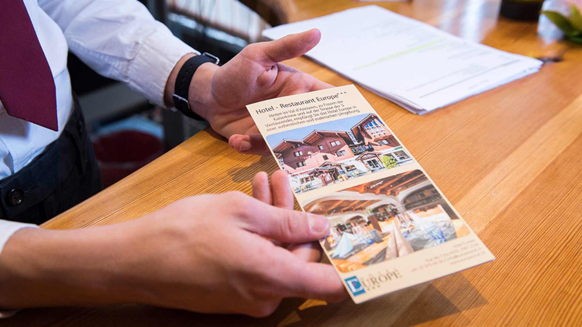 Une personne en chemise blanche et cravate rouge tient une brochure d'hôtel et de restaurant avec des images d'un bâtiment et de salles à manger sur une table en bois. Des papiers et une petite plante sont visibles à l'arrière-plan.