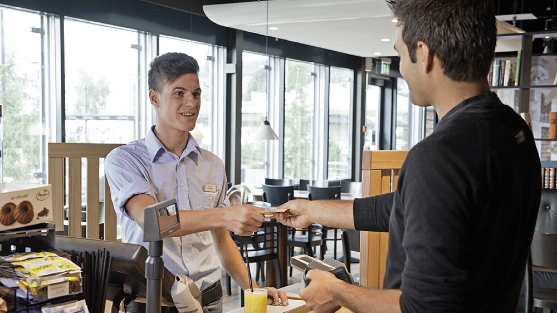 Un caissier souriant, vêtu d'une chemise bleu clair, remet un reçu à un client qui paie par carte de crédit au comptoir d'un magasin moderne où l'on peut voir des snacks et des boissons.