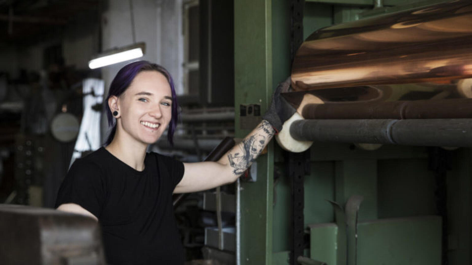 Une personne souriante aux cheveux violets et aux tatouages fait fonctionner des machines industrielles dans un atelier, vêtue d'une chemise et de gants noirs.