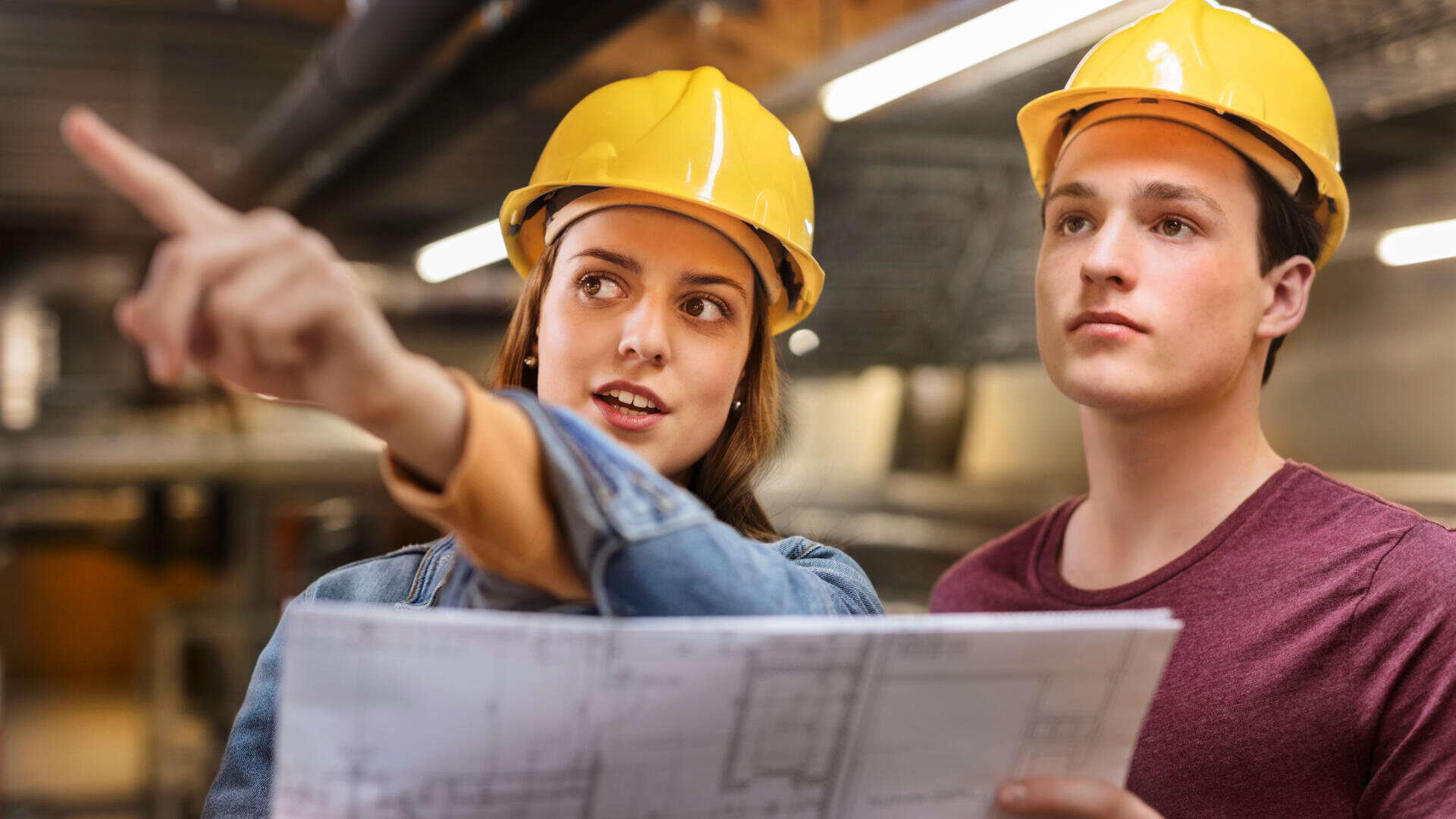 Deux personnes portant des casques jaunes se tiennent à l'intérieur ; l'une tient un plan en pointant au loin, l'autre regarde dans la même direction, toutes deux semblant concentrées sur un projet de construction ou d'architecture.