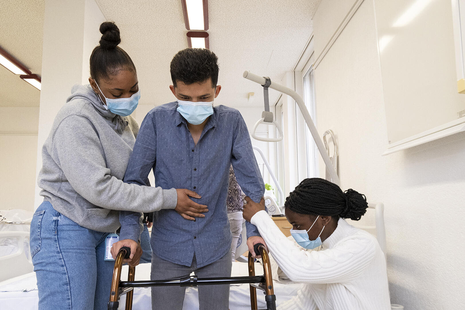 Trois personnes portant des masques se trouvent dans un environnement médical ; un homme s'appuie sur un déambulateur tandis que deux femmes l'assistent, l'une lui tenant le bras et l'autre guidant le déambulateur.