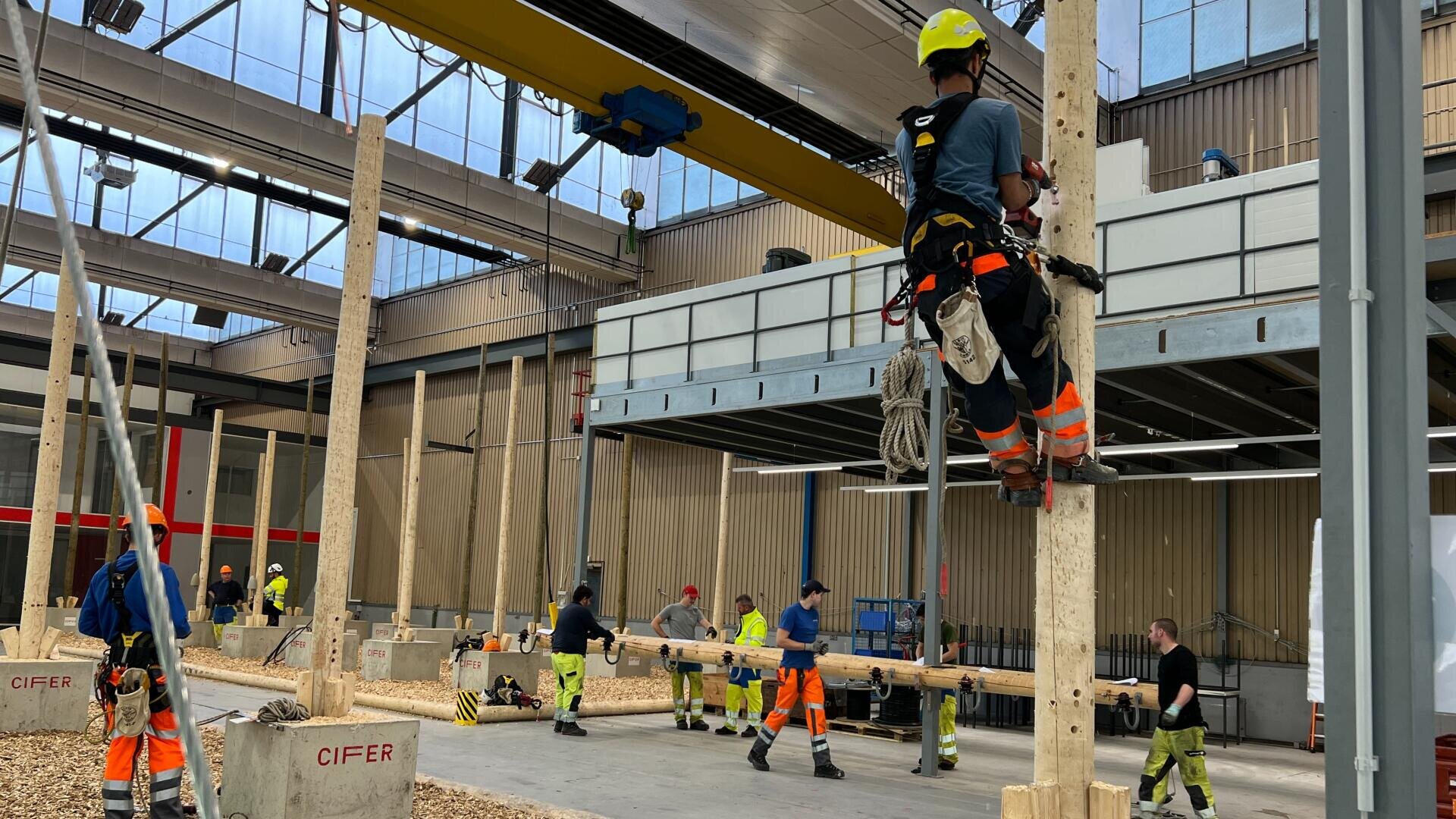 Des travailleurs portant des casques et des équipements de sécurité participent à un exercice de formation en intérieur, une personne grimpant sur un poteau en bois tandis que d'autres s'exercent ou observent dans une installation industrielle spacieuse.
