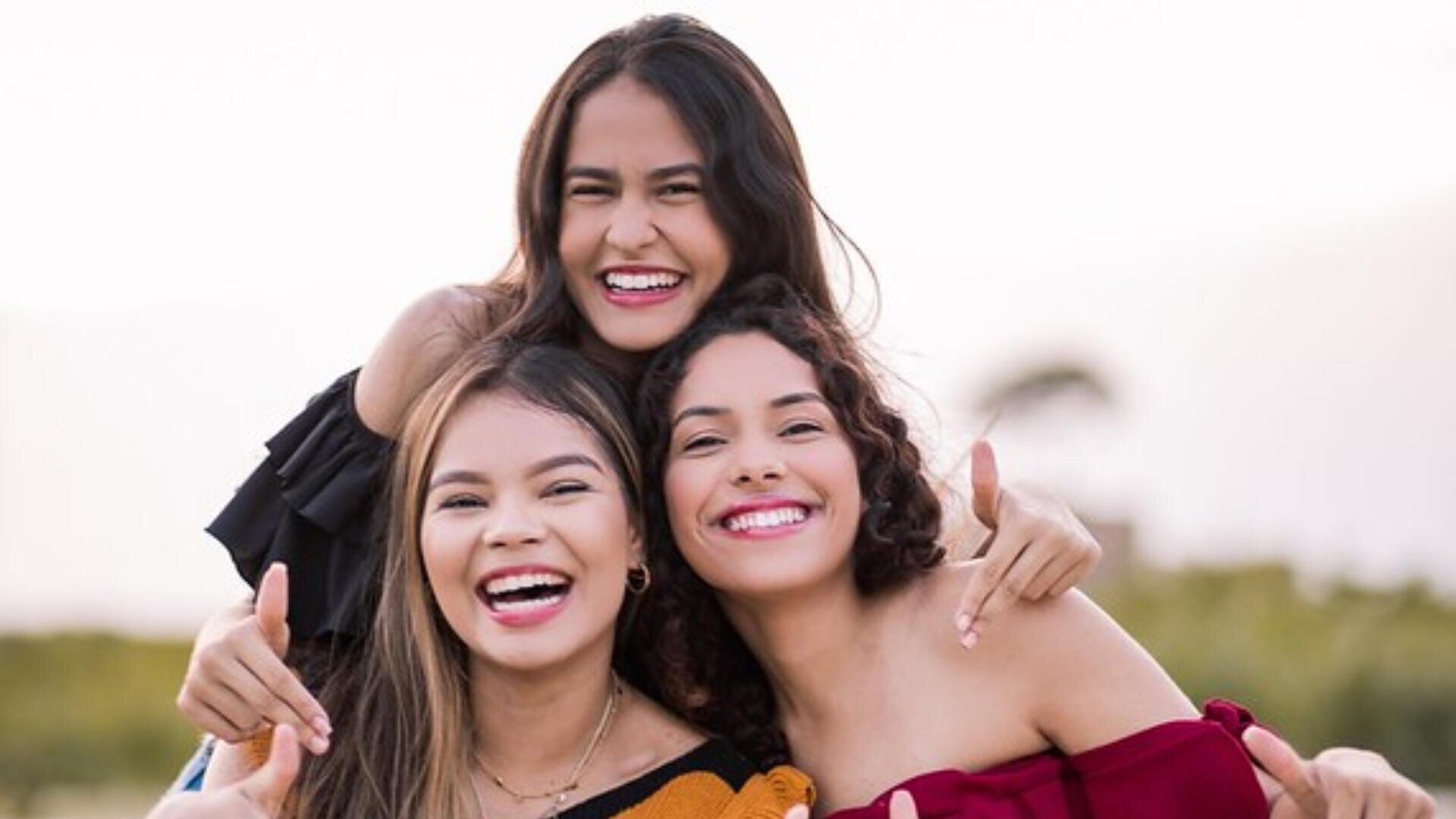Trois jeunes femmes se sourient et s'étreignent en plein air, avec de la verdure en arrière-plan. Elles semblent heureuses et joyeuses, profitant du temps qu'elles passent ensemble.
