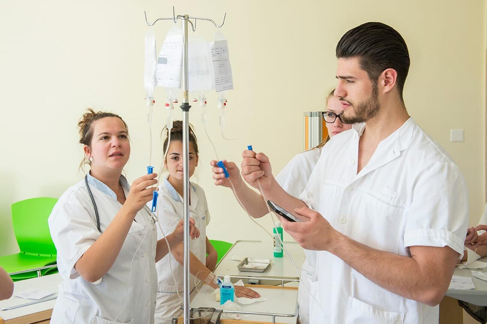 Quatre étudiants en médecine en blouse blanche travaillent ensemble à la mise en place de poches de perfusion et de tubulures dans une salle de classe clinique, tout en discutant et en prenant des notes.