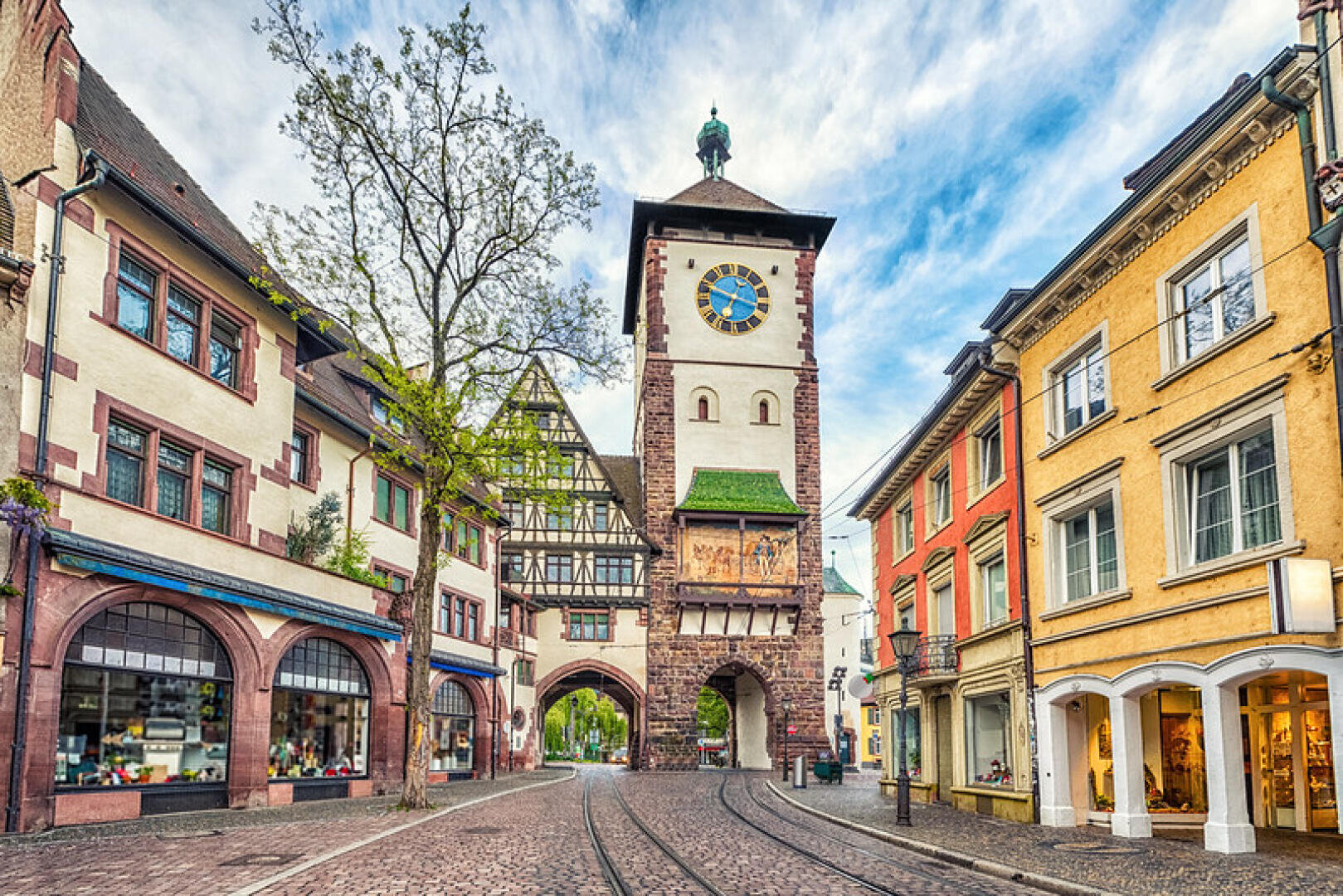 Une rue européenne pittoresque avec des bâtiments colorés, une tour d'horloge avec une porte voûtée, des rails de tramway sur une chaussée pavée, et un ciel partiellement nuageux au-dessus de la tête.