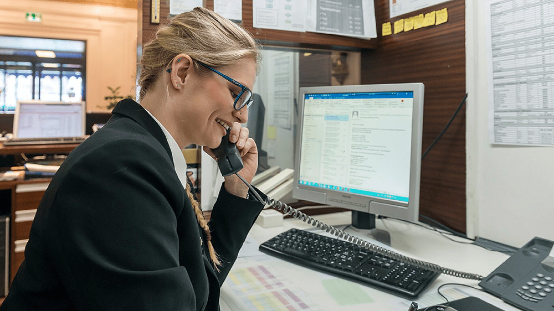 Une femme en tenue de travail sourit tout en parlant au téléphone à son bureau, en regardant un écran d'ordinateur avec des courriels ouverts. L'espace de travail est organisé avec des documents, un clavier, une souris et des fournitures de bureau.