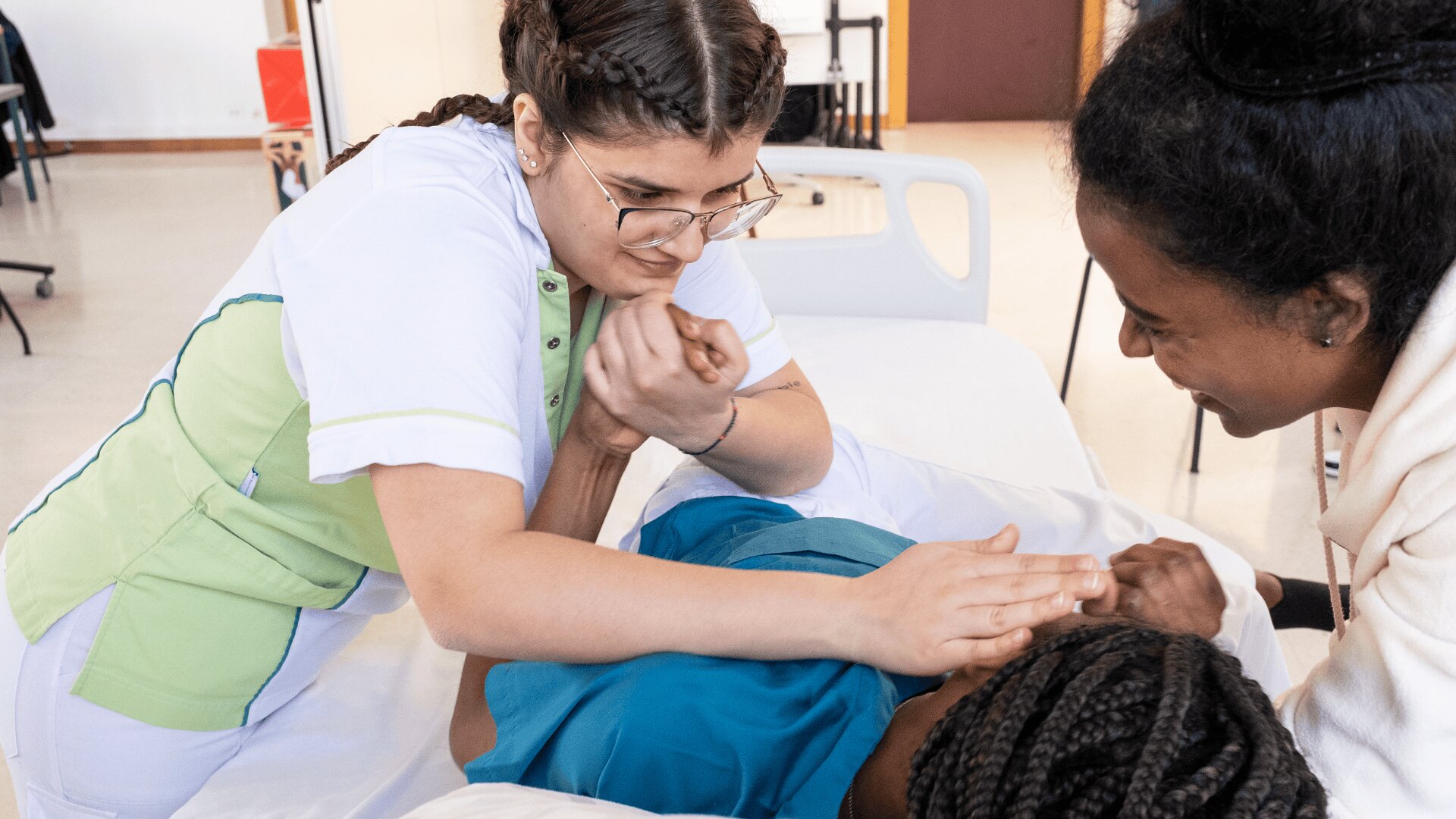 Un agent de santé aide une personne allongée dans un lit d'hôpital, en la soutenant délicatement avec les mains, tandis qu'une autre personne la regarde en souriant.