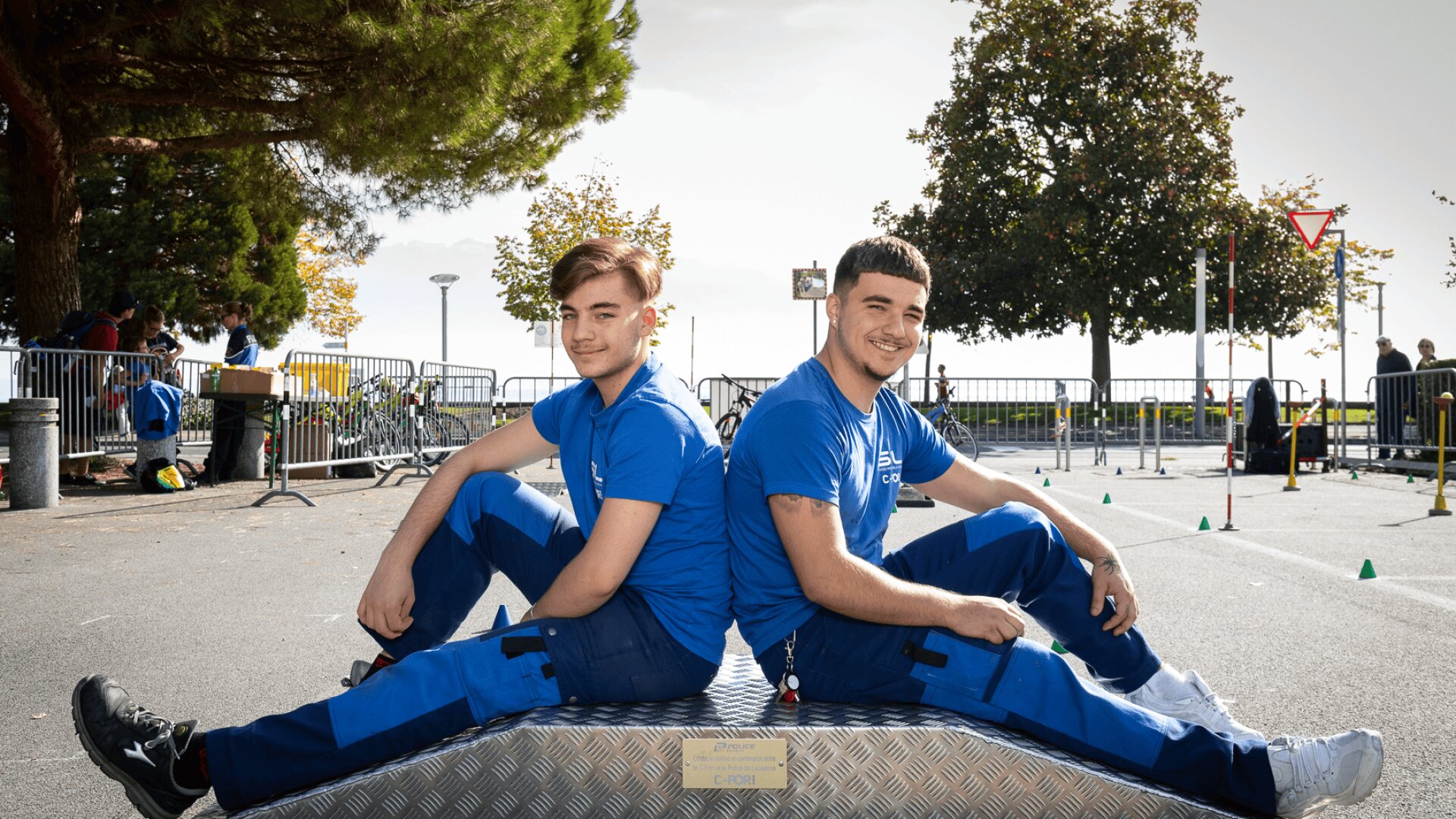 Deux jeunes hommes portant des uniformes bleus sont assis dos à dos sur une plate-forme métallique en plein air et sourient à la caméra. Des arbres, des clôtures et des personnes sont visibles à l'arrière-plan par une journée ensoleillée.