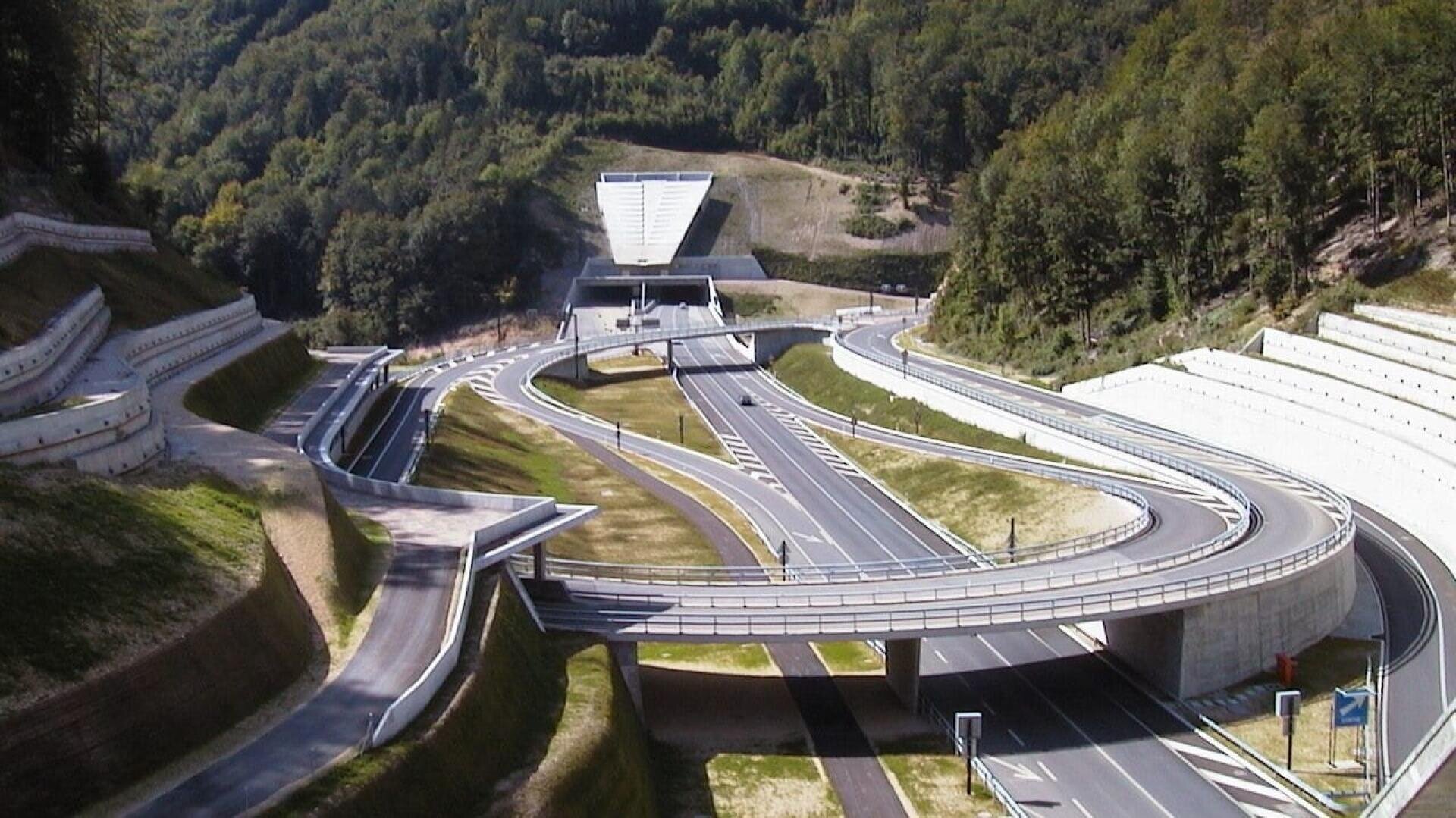 Un échangeur autoroutier complexe avec des routes sinueuses et des rampes d'accès dans une région vallonnée et boisée, menant à l'entrée d'un tunnel à l'arrière-plan. Des barrières en béton et des marquages routiers sont visibles tout au long de la scène.