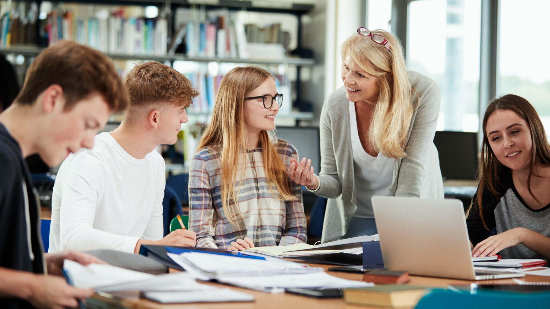 Dans une salle de classe ou une bibliothèque moderne et lumineuse, un enseignant sourit et discute avec quatre élèves assis à une table couverte de livres, de documents et d'un ordinateur portable. Les élèves sont engagés et écoutent attentivement.
