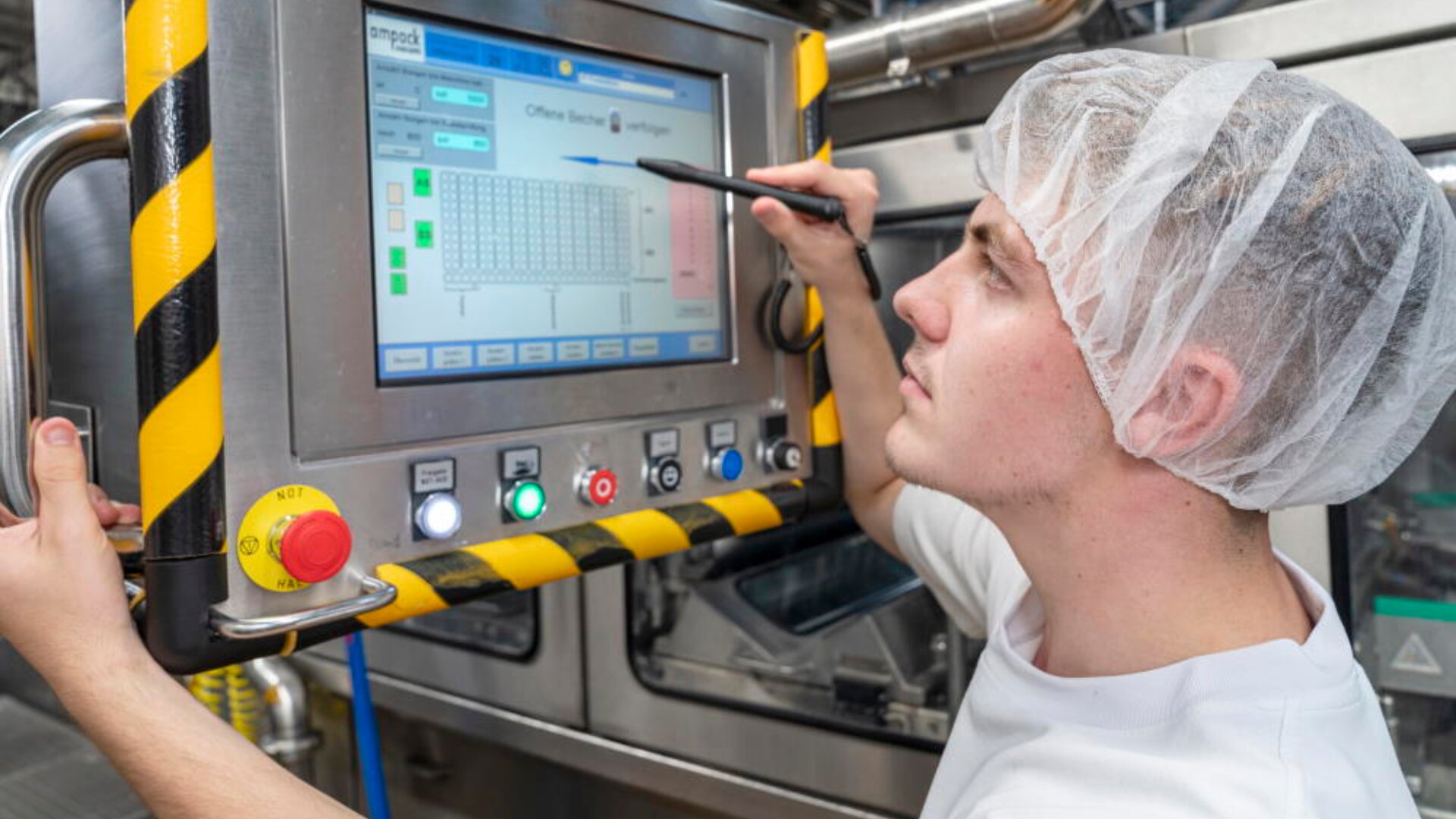 Un travailleur portant une charlotte utilise un stylet pour faire fonctionner un panneau de commande tactile dans un environnement industriel, entouré de machines et de bordures de sécurité.