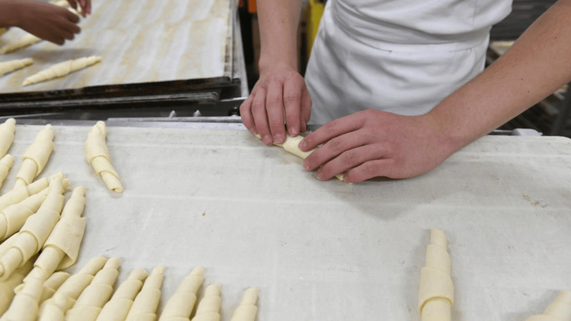 Une personne vêtue d'un tablier blanc roule la pâte en forme de croissant sur une surface farinée, tandis que plusieurs croissants non cuits sont alignés à proximité.