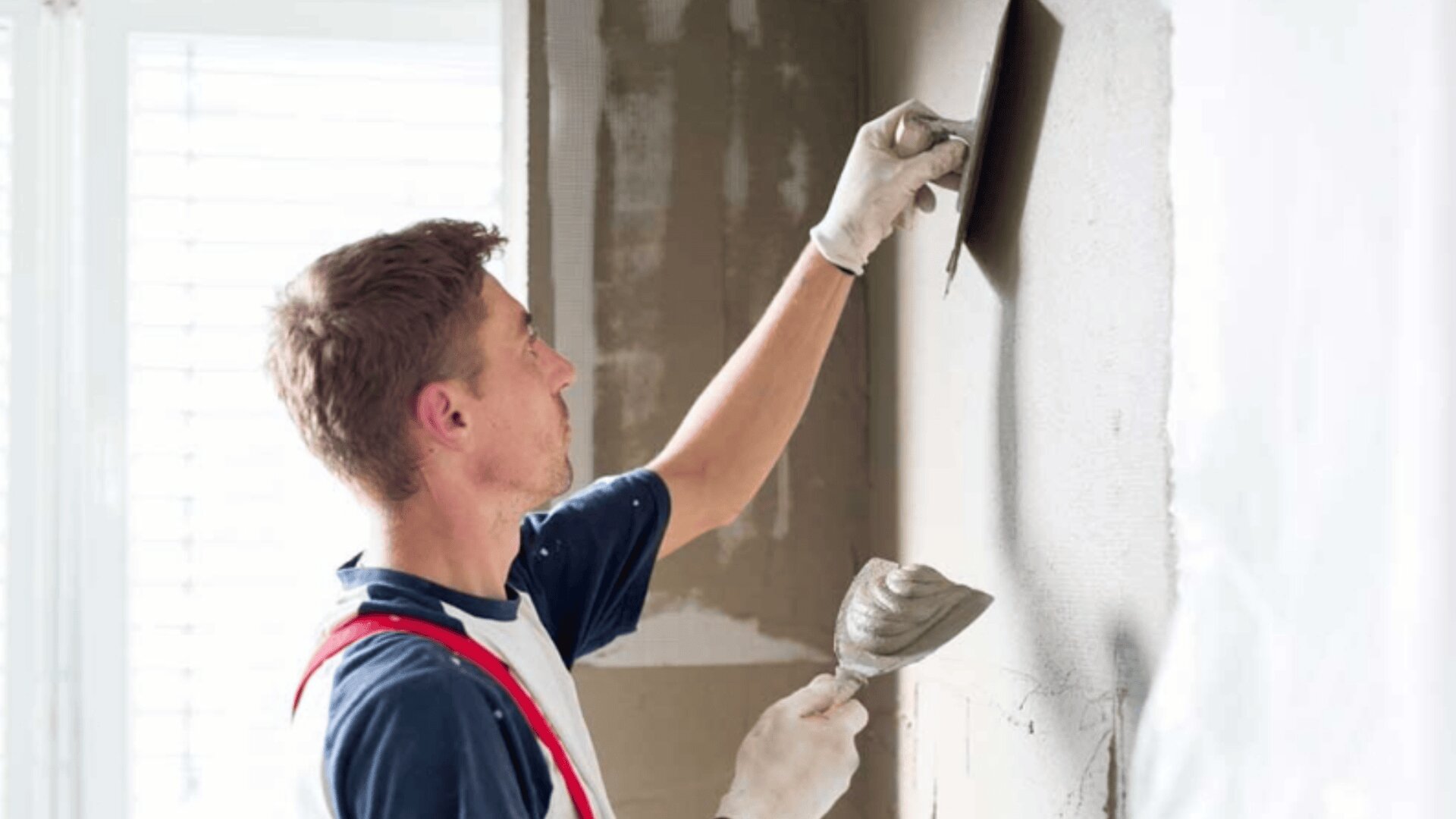 Un homme en tenue de travail et ganté utilise une truelle pour appliquer du plâtre sur un mur intérieur. Il tient dans l'autre main un faucon avec du plâtre. La pièce est bien éclairée par la lumière naturelle d'une fenêtre.
