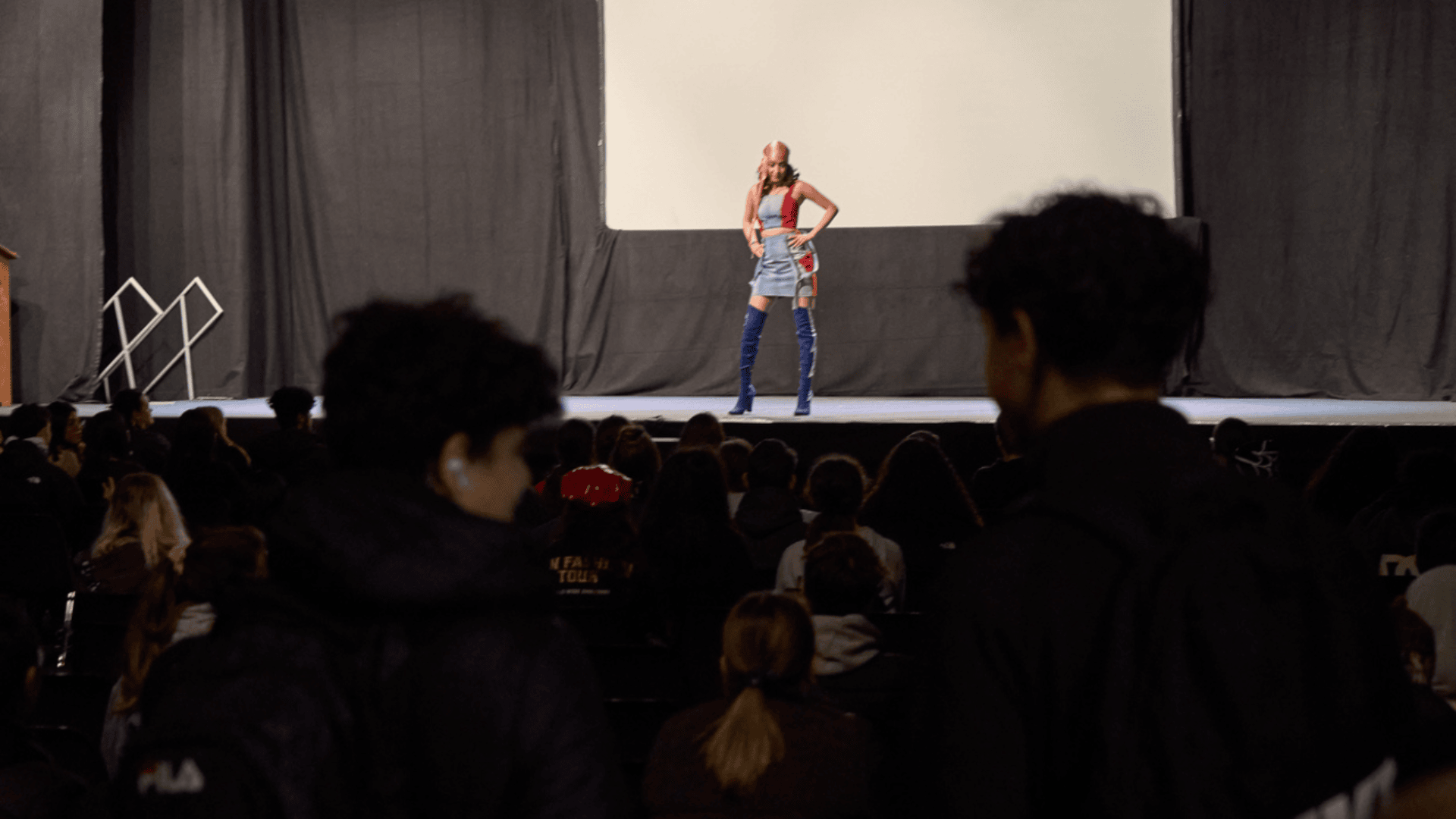 Un artiste vêtu d'une tenue colorée et de bottes bleues se tient sur scène devant un public assis, avec un grand écran vide derrière lui et des rideaux sombres de part et d'autre.