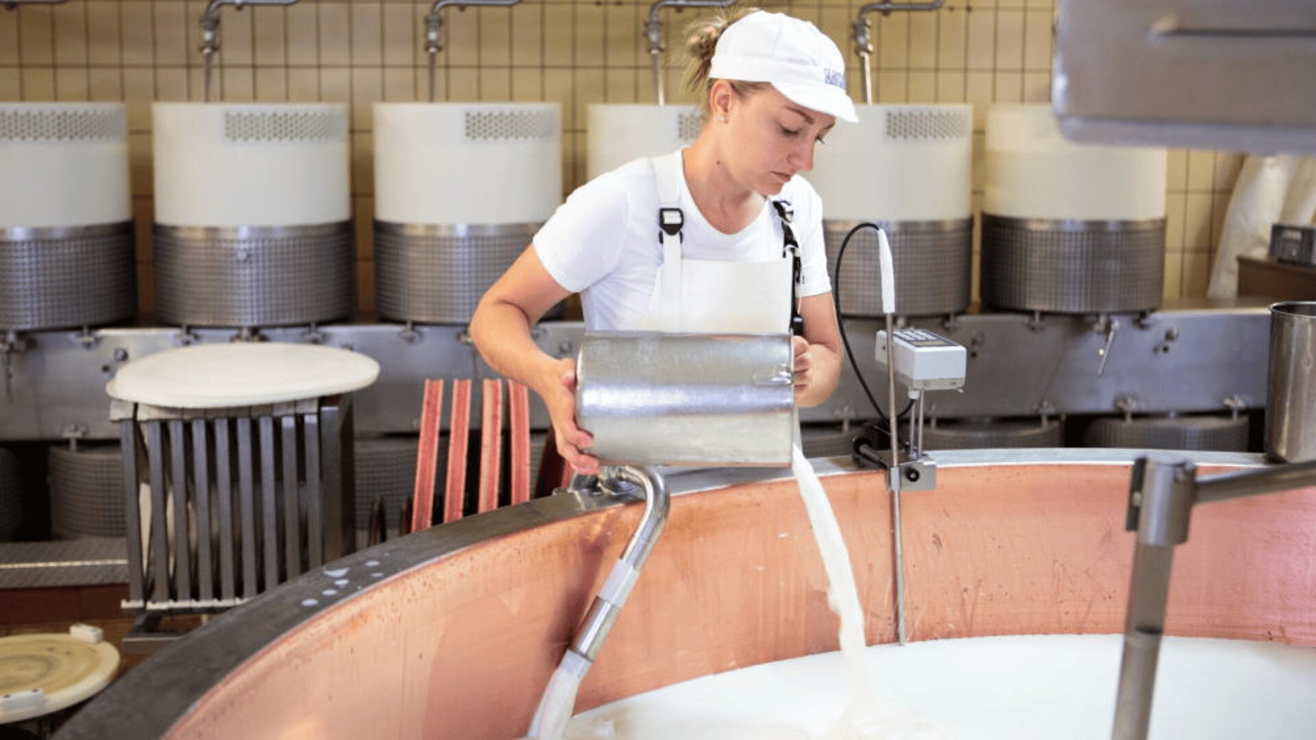 Une personne en tenue de travail blanche et coiffée d'une casquette verse un liquide d'un récipient métallique dans une grande cuve en cuivre dans une laiterie ou une fromagerie, avec des équipements à l'arrière-plan.