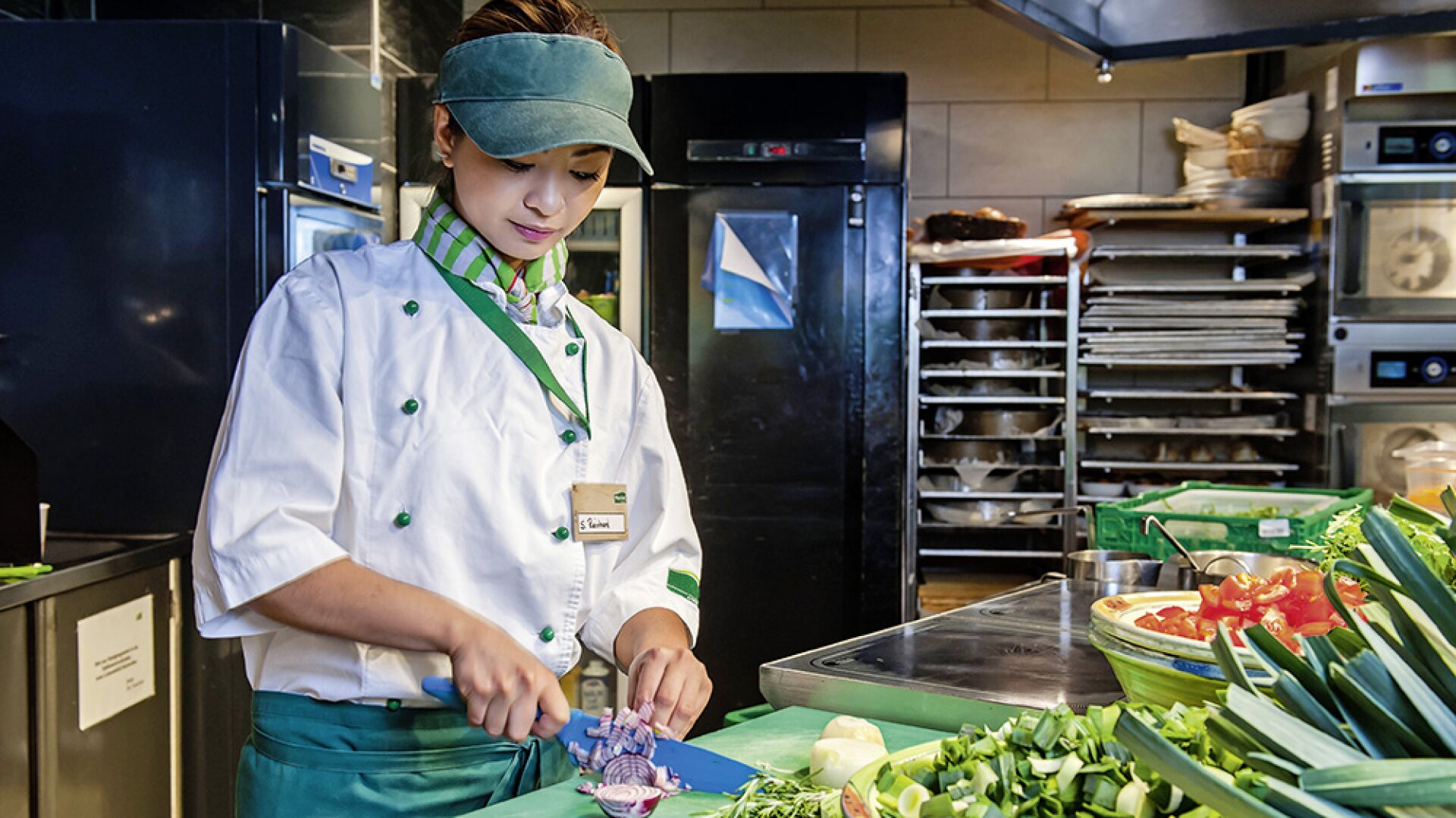 Un chef portant un uniforme blanc, un tablier vert et une visière coupe des légumes dans une cuisine commerciale, entouré de produits frais et de matériel de cuisson.