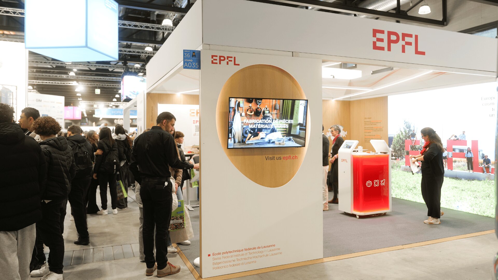 Stand d'exposition de l'EPFL lors d'un événement en intérieur, avec un écran d'affichage, un comptoir d'information et des personnes qui interagissent avec les objets exposés. Le stand est moderne, avec la marque de l'EPFL visible sur les murs et les présentoirs.
