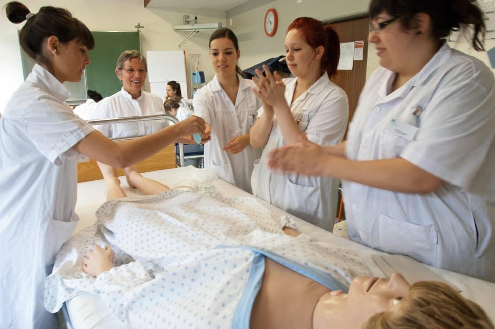 Cinq professionnels de la santé en uniforme blanc se rassemblent autour d'un lit d'hôpital, pratiquant une procédure sur un mannequin de formation médicale dans une salle de classe clinique. Certains sourient et préparent du matériel médical.