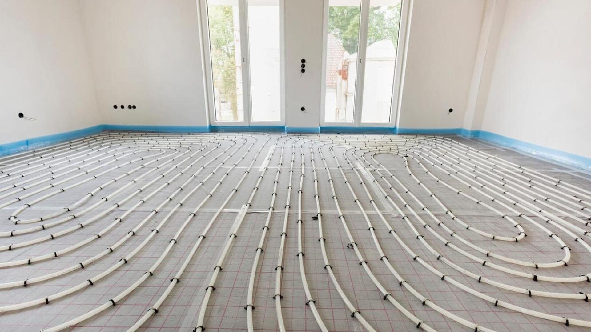 Une pièce vide et lumineuse, dotée de grandes fenêtres, présente un système de chauffage par le sol récemment installé, avec des tuyaux blancs disposés en grille sur le sol.