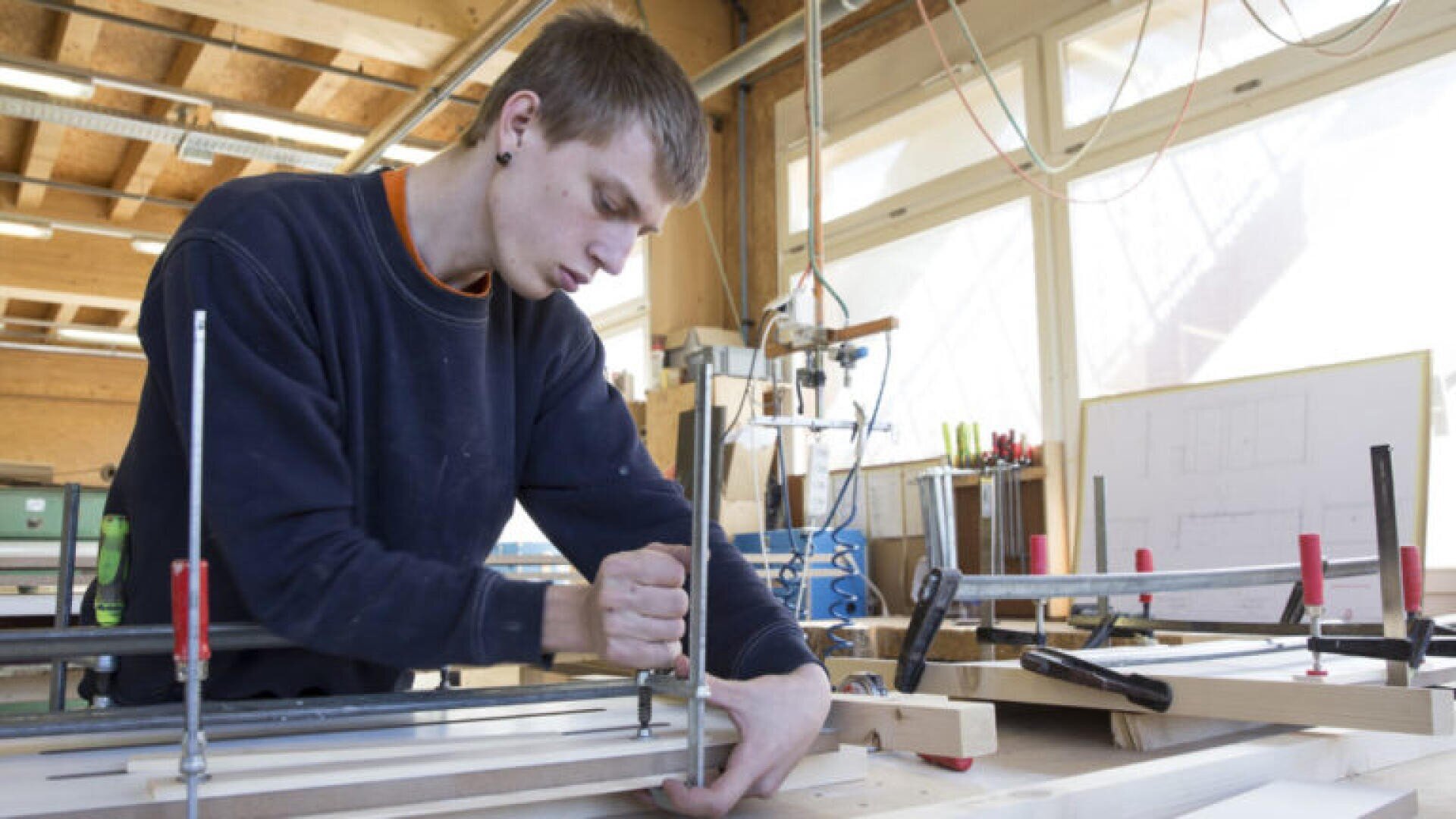 Dans un atelier, un jeune utilise un tournevis sur un projet en bois, en serrant les pièces ensemble sur un établi. L'espace de travail est bien éclairé par de grandes fenêtres et des outils sont accrochés au mur à l'arrière-plan.