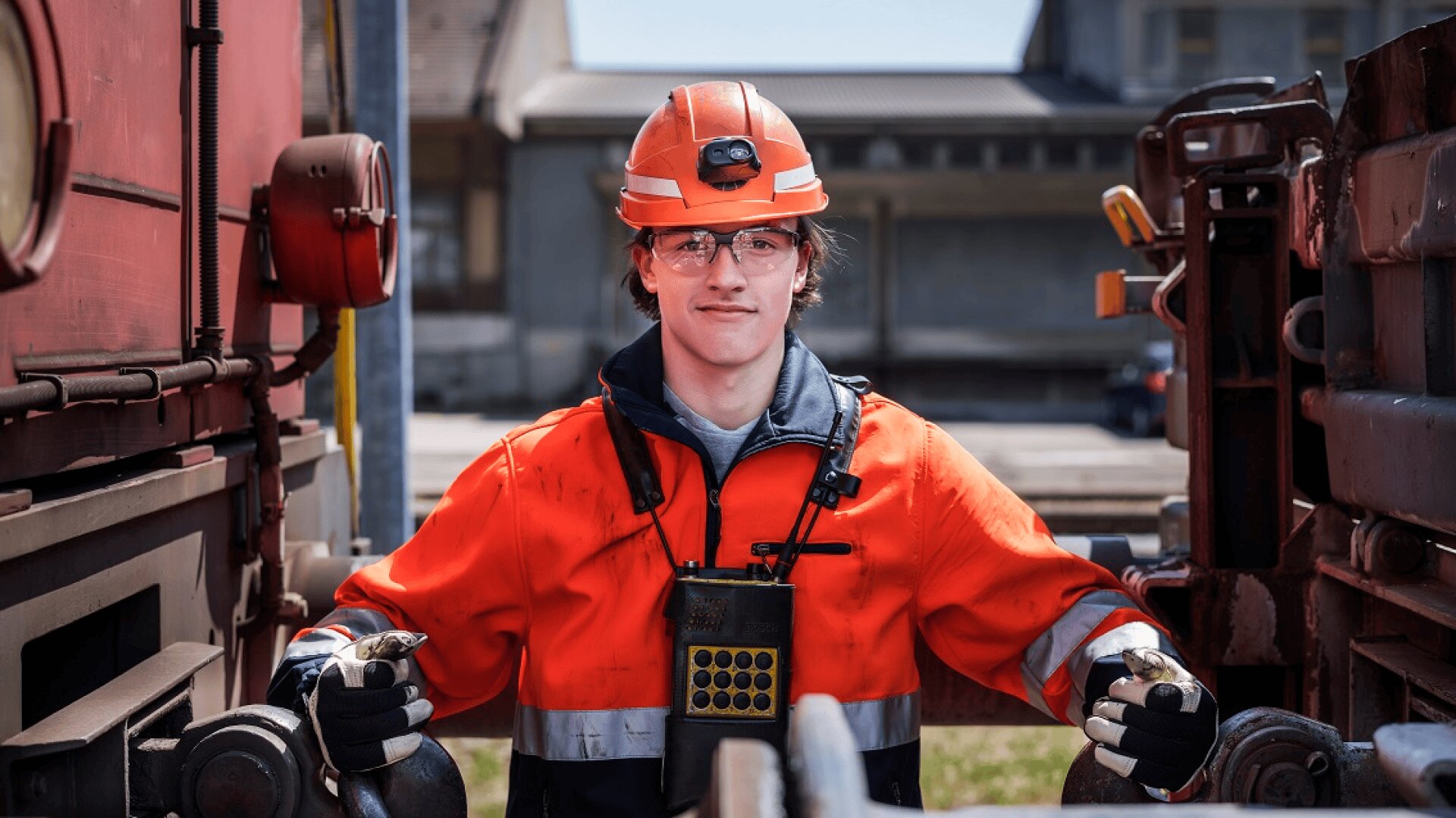 Un cheminot portant un uniforme de sécurité orange, un casque et des lunettes se tient entre deux wagons de train, actionnant un équipement d'attelage à l'extérieur par une journée ensoleillée.