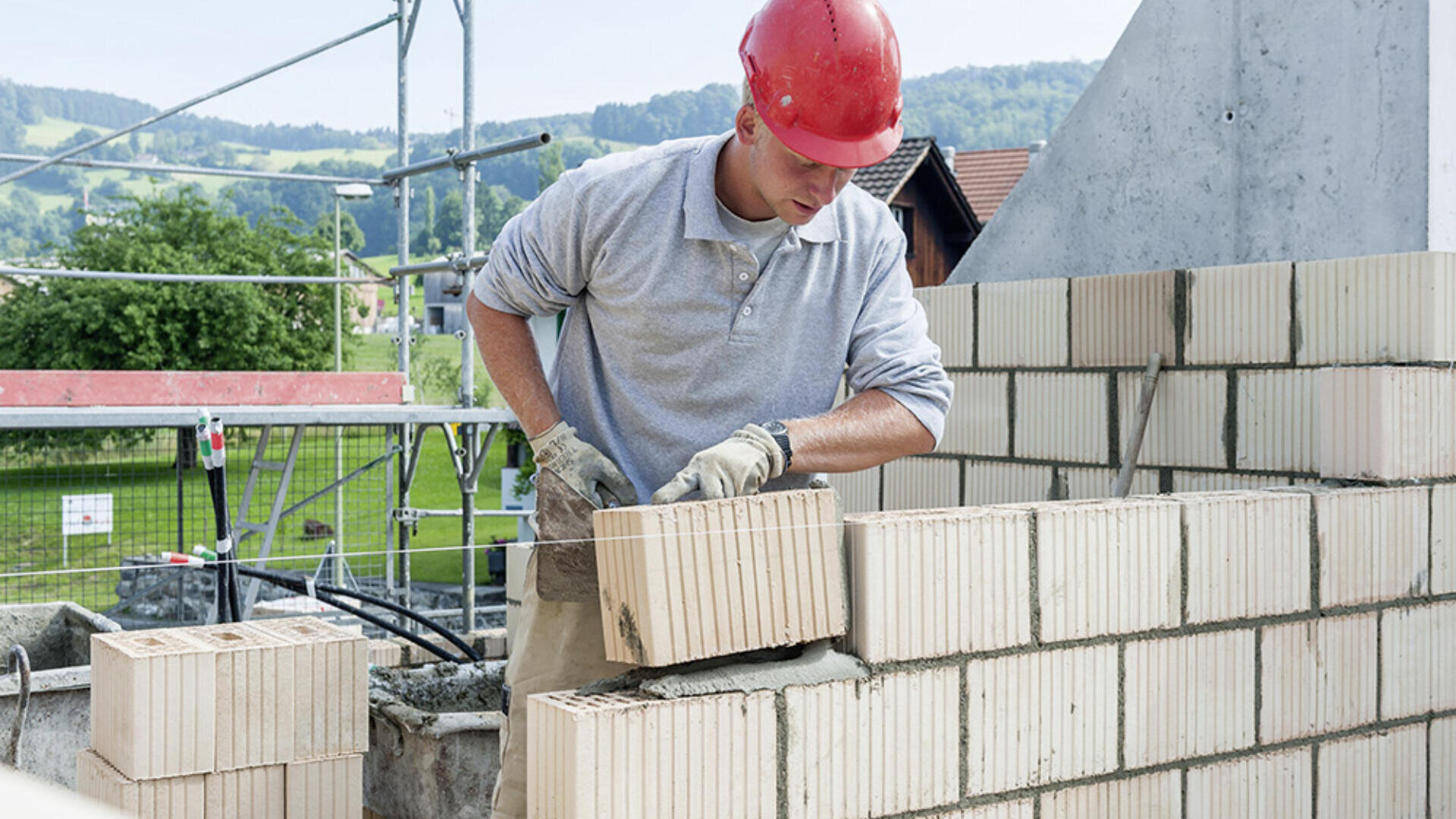 Un ouvrier du bâtiment portant un casque rouge et une chemise grise pose soigneusement une grosse brique sur un mur sur un chantier extérieur avec des échafaudages et de la verdure en arrière-plan.