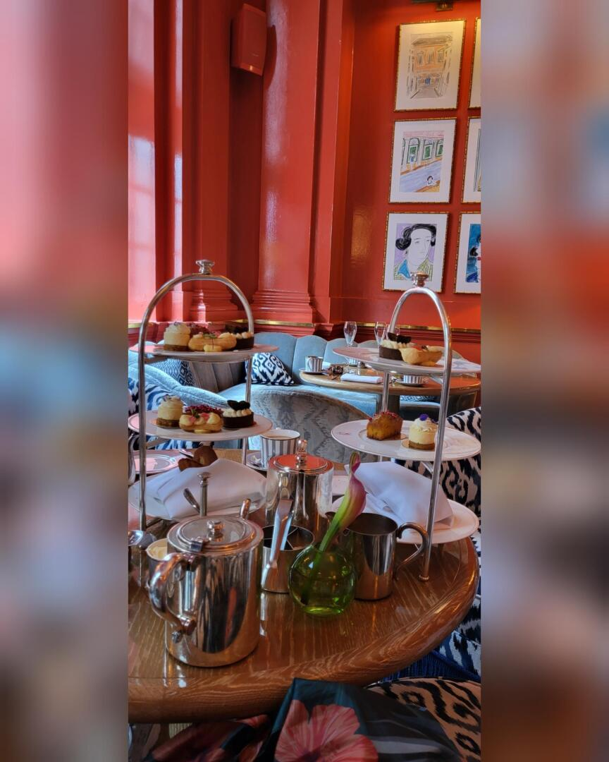 Une table dressée pour le thé de l'après-midi avec des plateaux étagés de pâtisseries, une théière et des verres dans une pièce lumineuse aux murs orangés avec des œuvres d'art encadrées en arrière-plan.