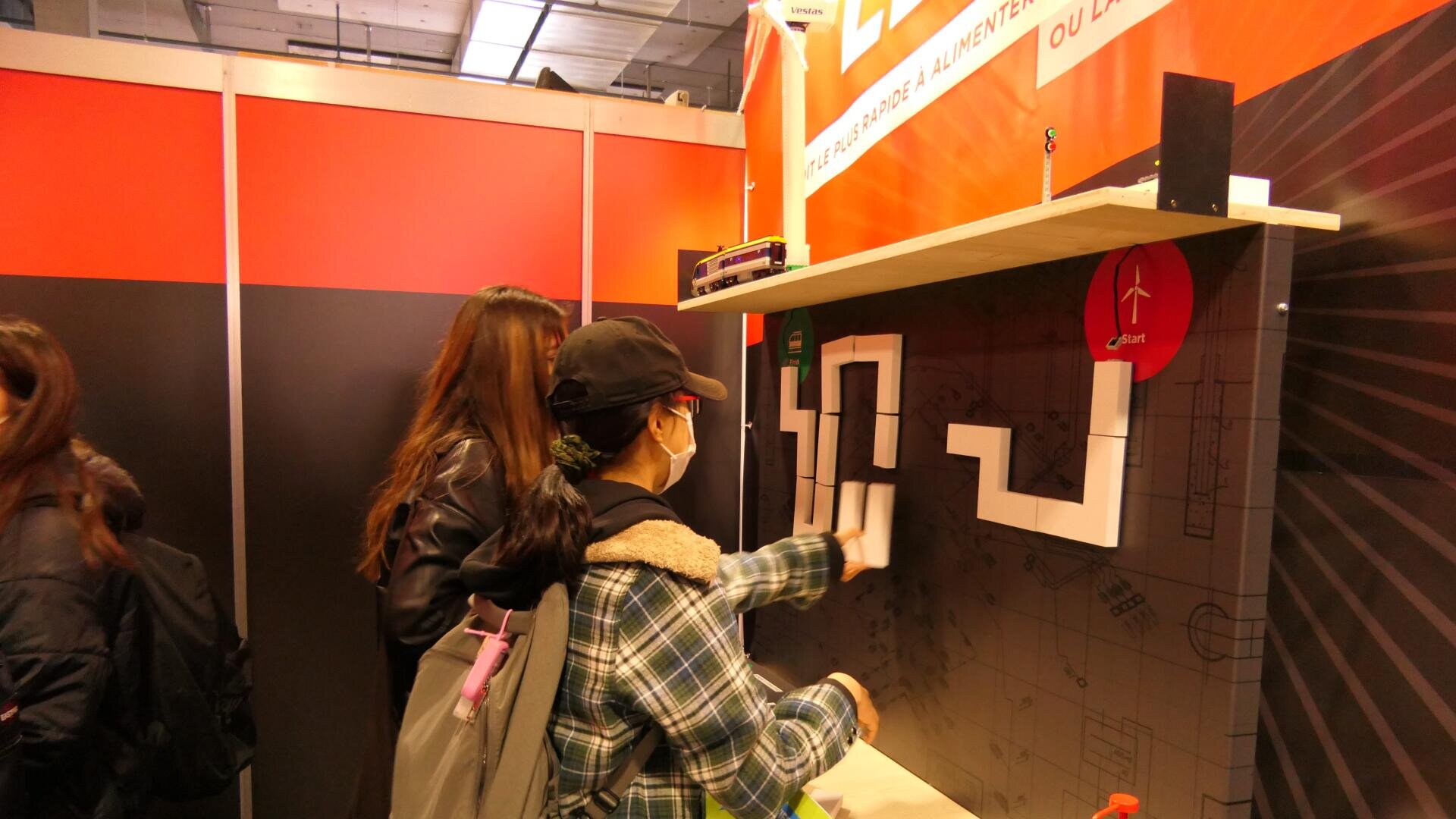 Plusieurs personnes interagissent avec un jeu de puzzle mural lors d'un événement ou d'un stand d'exposition en intérieur, en déplaçant des blocs blancs pour les faire entrer dans les contours noirs de l'écran.
