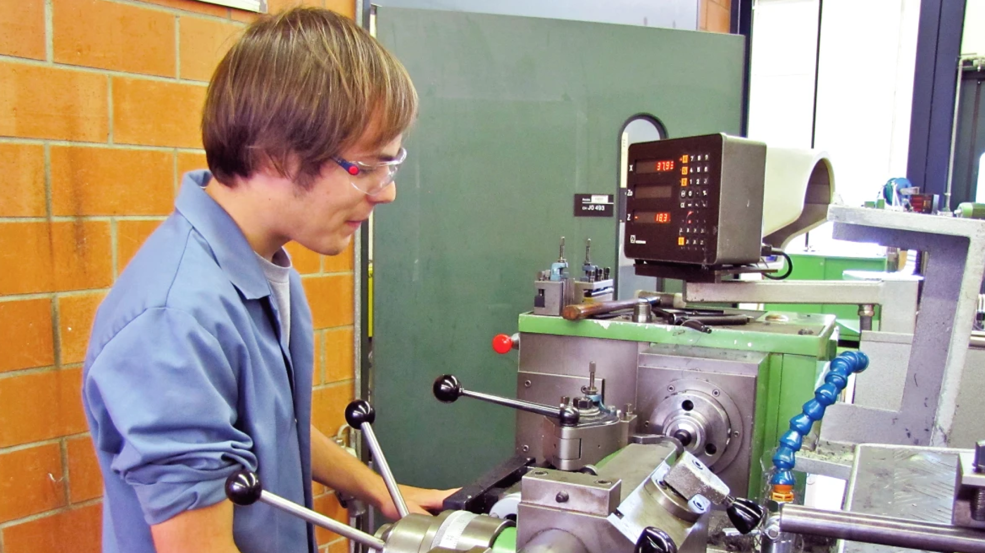 Une personne portant des lunettes de sécurité utilise un tour dans un atelier. Le tour est équipé d'un écran de lecture numérique et divers outils sont visibles sur la machine et la table voisine.