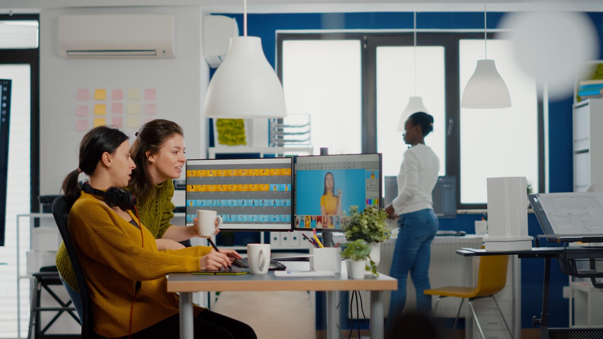 Deux femmes sont assises à un bureau et travaillent sur des ordinateurs dont les écrans affichent des palettes de couleurs et des photos, tandis qu'une autre femme se tient à l'arrière-plan dans un bureau moderne et bien éclairé.