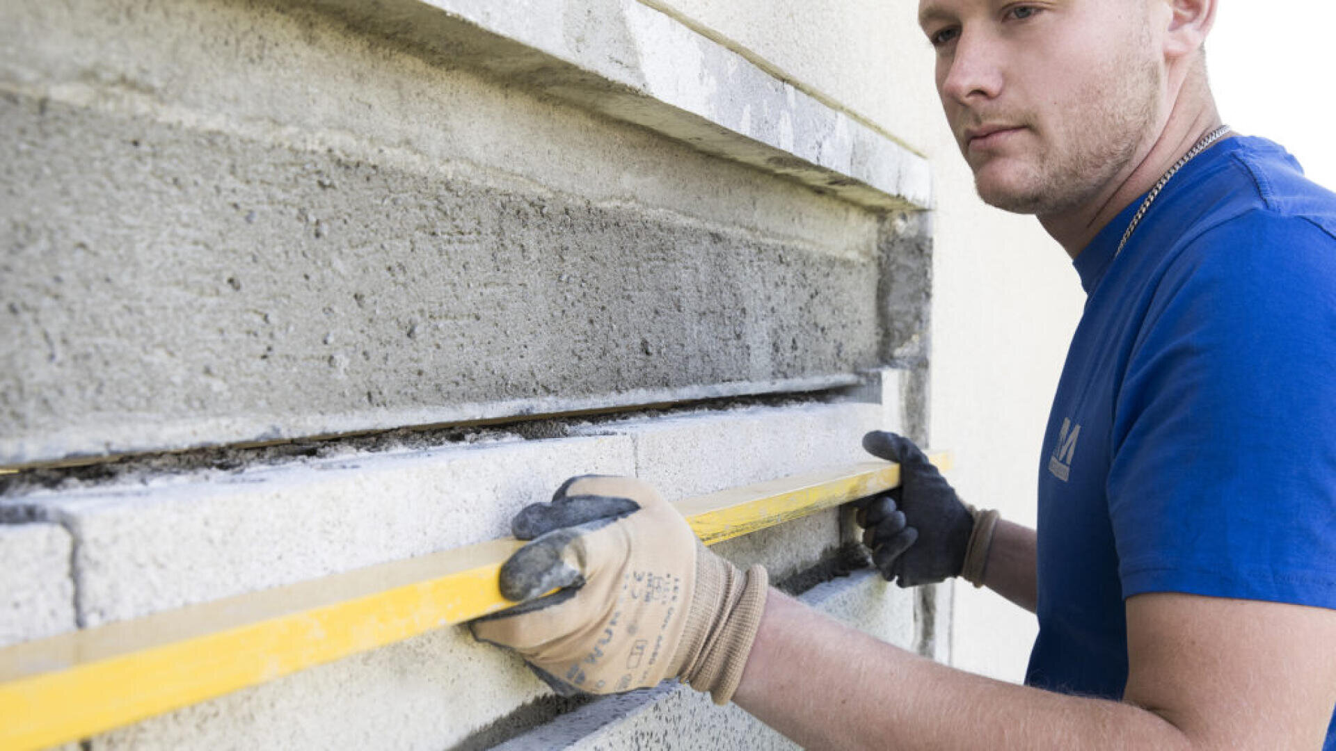 Un ouvrier du bâtiment portant des gants utilise un niveau à bulle jaune pour vérifier l'alignement des blocs de béton sur un mur extérieur.