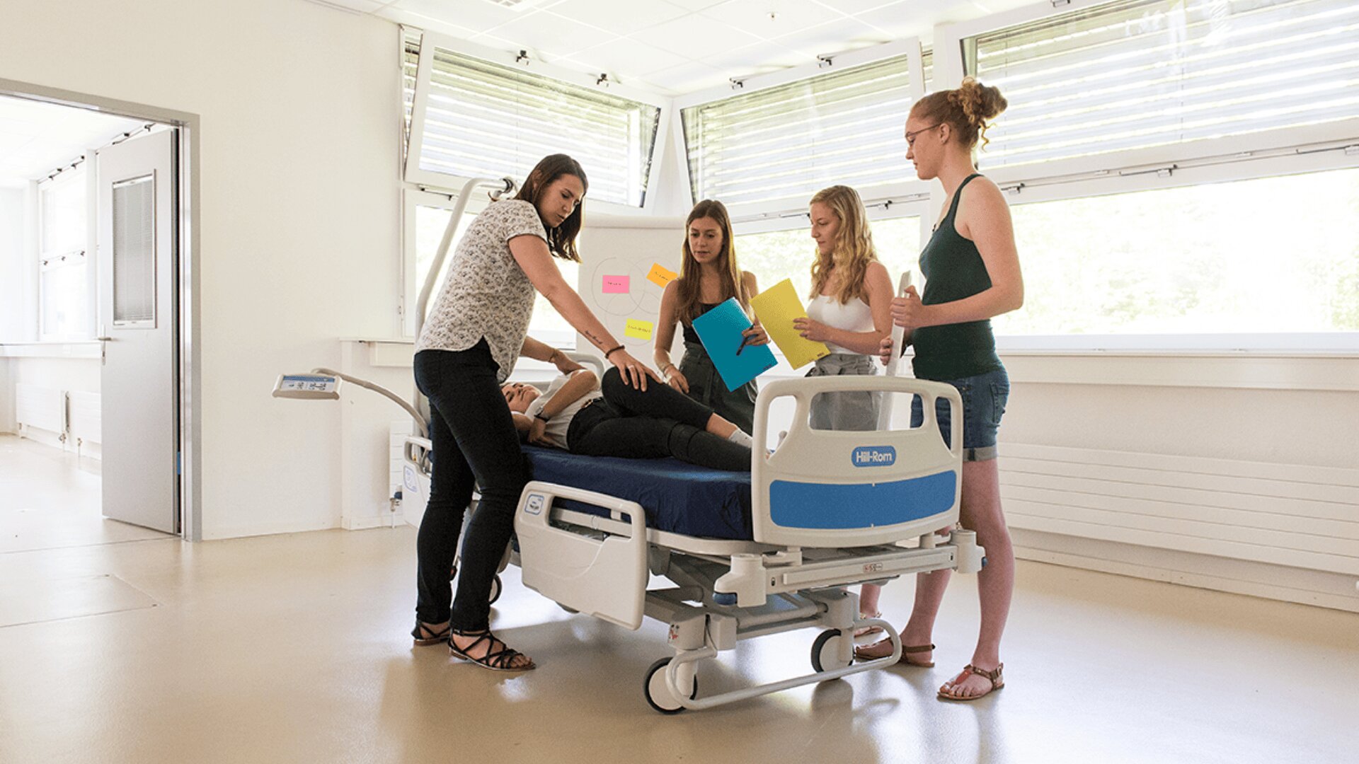 Quatre femmes se tiennent autour d'un lit d'hôpital dans une pièce lumineuse, l'une d'entre elles étant allongée sur le lit. Le groupe semble engagé dans une discussion ou une session de formation, avec des notes sur un tableau à l'arrière-plan.