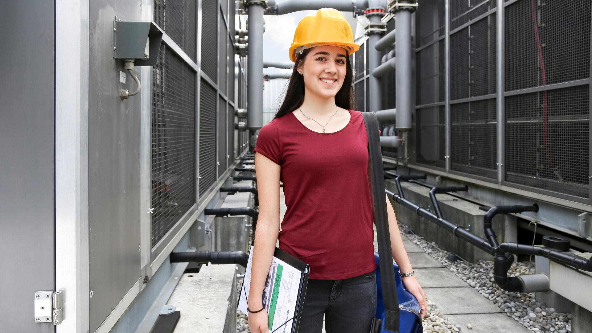 Une femme portant un casque jaune, une chemise rouge et des documents se tient entre des tuyaux et des équipements industriels, souriant à la caméra.
