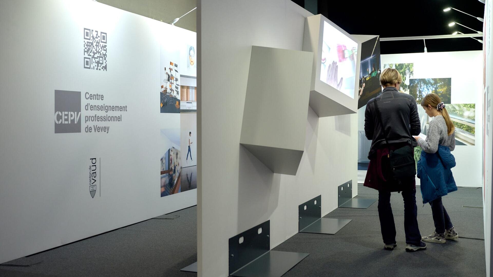 Deux personnes se tiennent près d'un présentoir d'exposition blanc avec des images, des codes QR et des logos, dont "Centre d'enseignement professionnel de Vevey". L'espace est doté d'étagères et d'éclairages modernes et minimalistes.