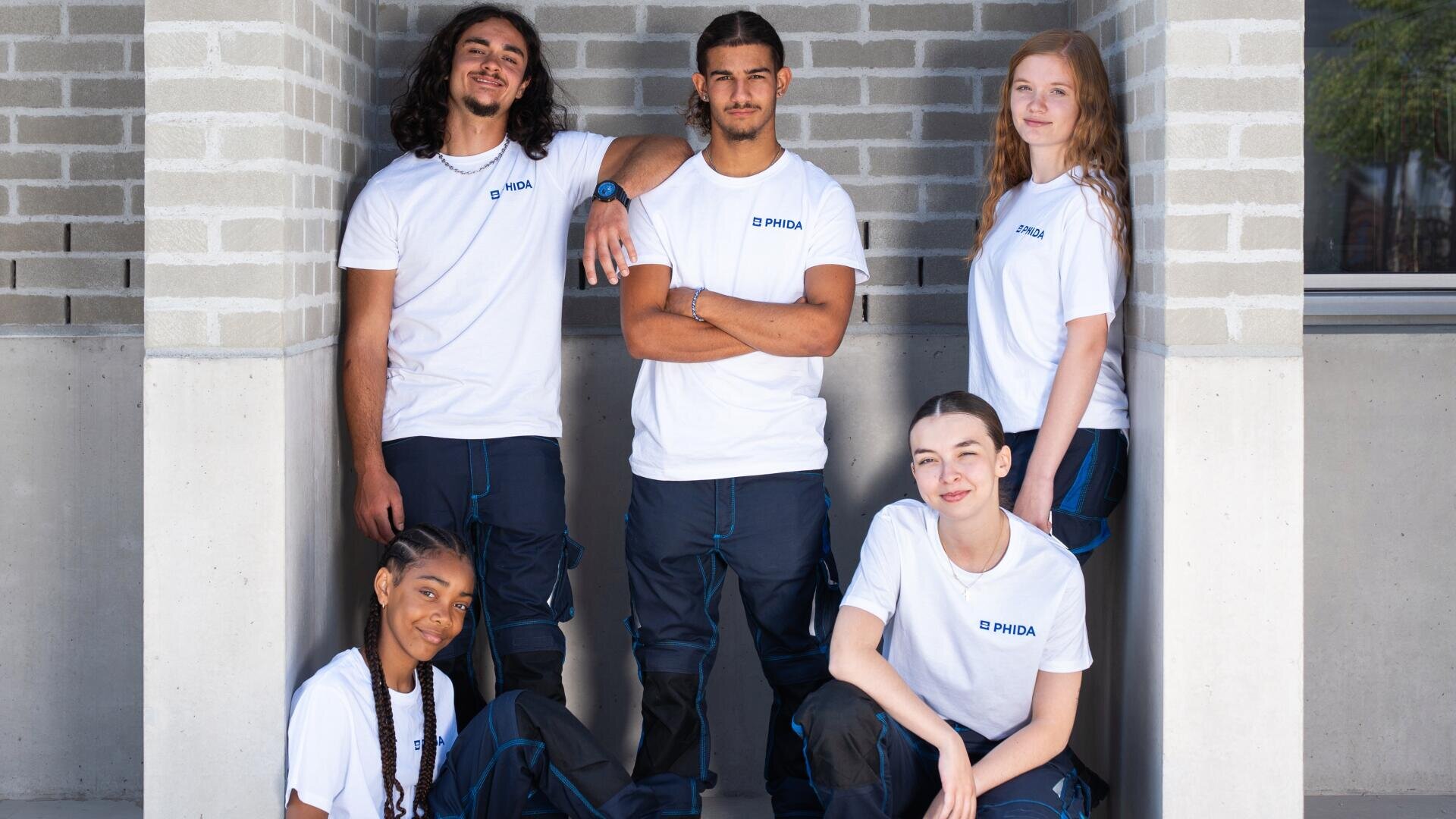 Cinq jeunes adultes portant des t-shirts blancs et des pantalons sombres assortis posent ensemble contre un mur de briques. Ils semblent détendus et confiants, certains étant debout et d'autres assis.