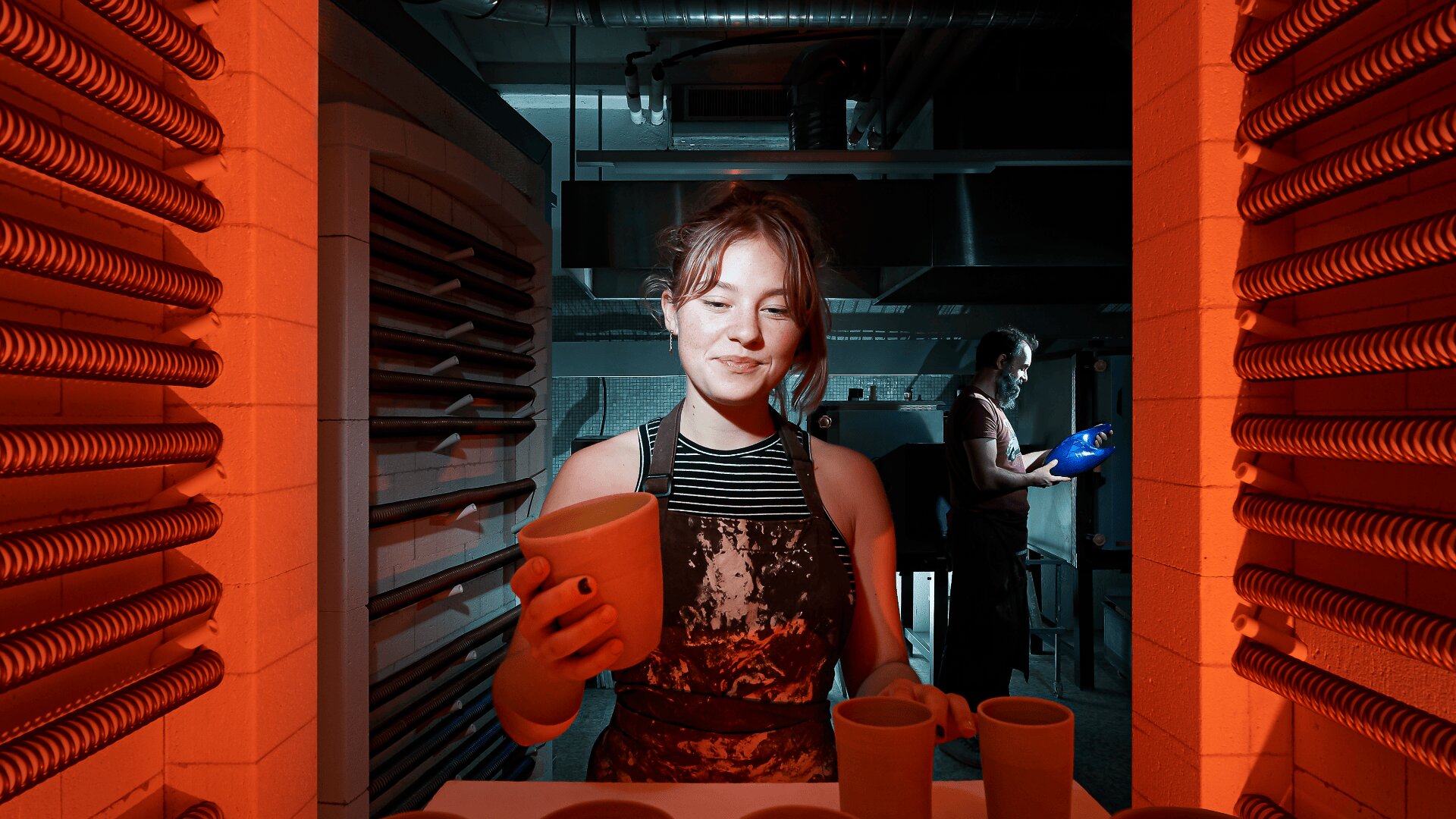 Une femme en tablier sourit en tenant un pot d'argile à l'intérieur d'un atelier de poterie sous un éclairage orange ; à l'arrière-plan, un homme travaille près d'un four sous une lumière bleue.