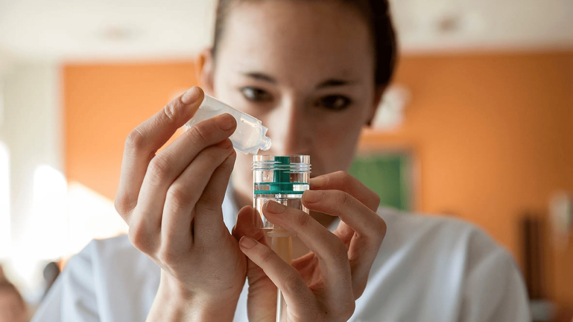 Une personne en blouse blanche verse avec précaution un liquide d'un flacon compte-gouttes dans un petit récipient transparent, en se concentrant sur sa tâche. L'arrière-plan est légèrement flou, avec des murs aux tons chauds.