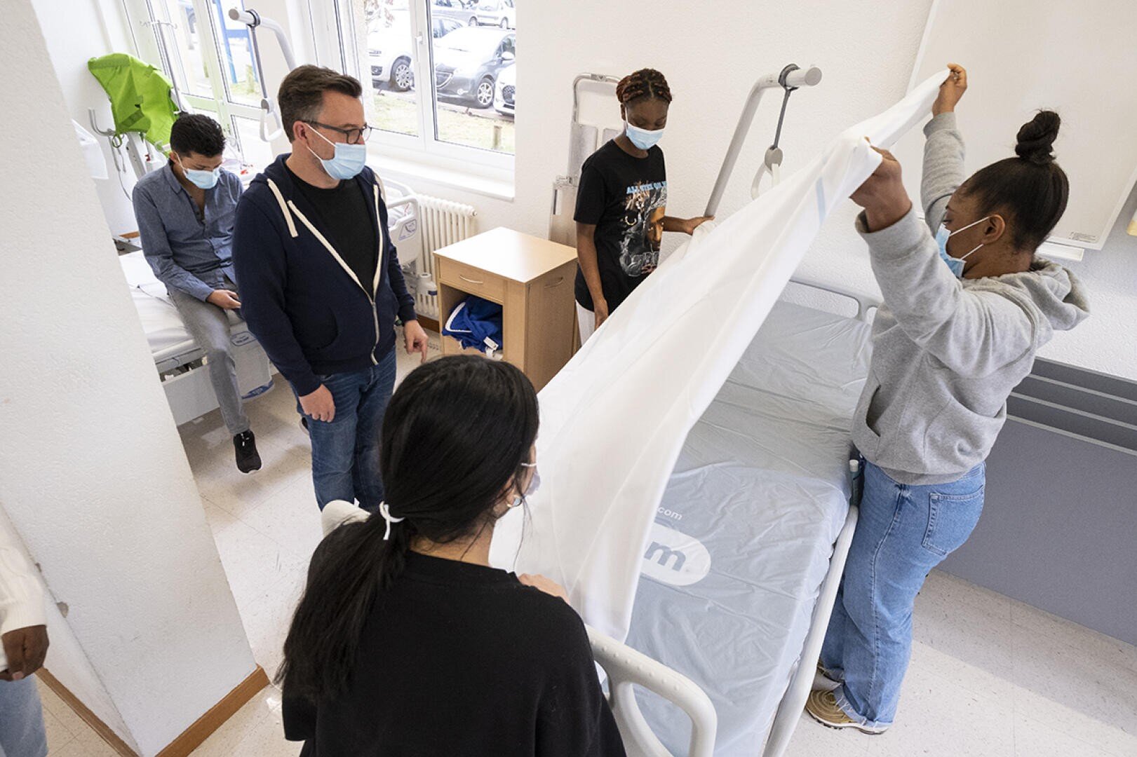 Plusieurs personnes portant des masques sont réunies dans une chambre d'hôpital. Deux personnes font un lit tandis que d'autres observent. La pièce est équipée de matériel médical et d'une fenêtre laissant passer la lumière du jour.