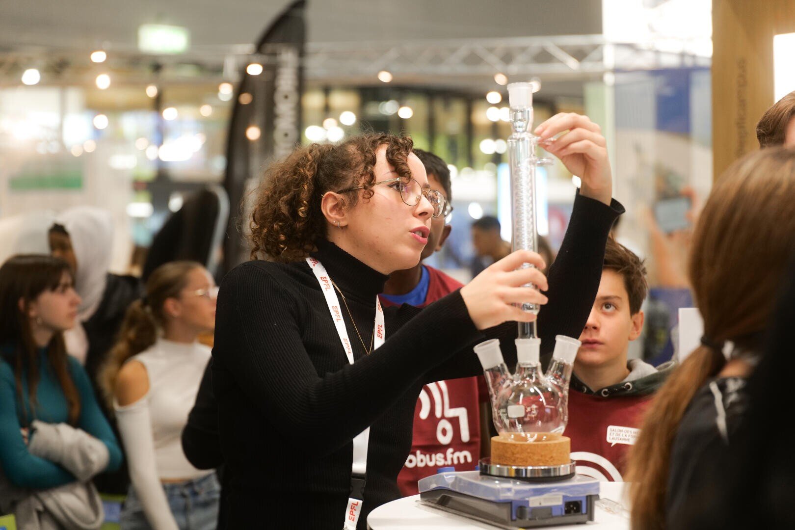 Une jeune femme portant des lunettes démontre une expérience scientifique avec du matériel de laboratoire à un groupe d'étudiants attentifs lors d'un événement ou d'une exposition en intérieur.