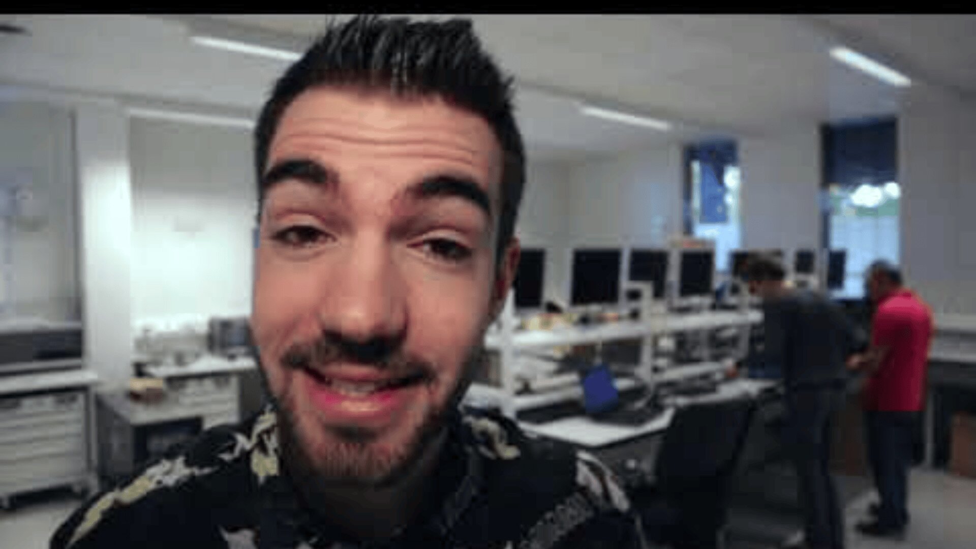 Un homme aux cheveux noirs et à la barbe bien taillée sourit à la caméra dans un bureau ou un laboratoire moderne et lumineux, avec en arrière-plan deux personnes travaillant sur des ordinateurs.