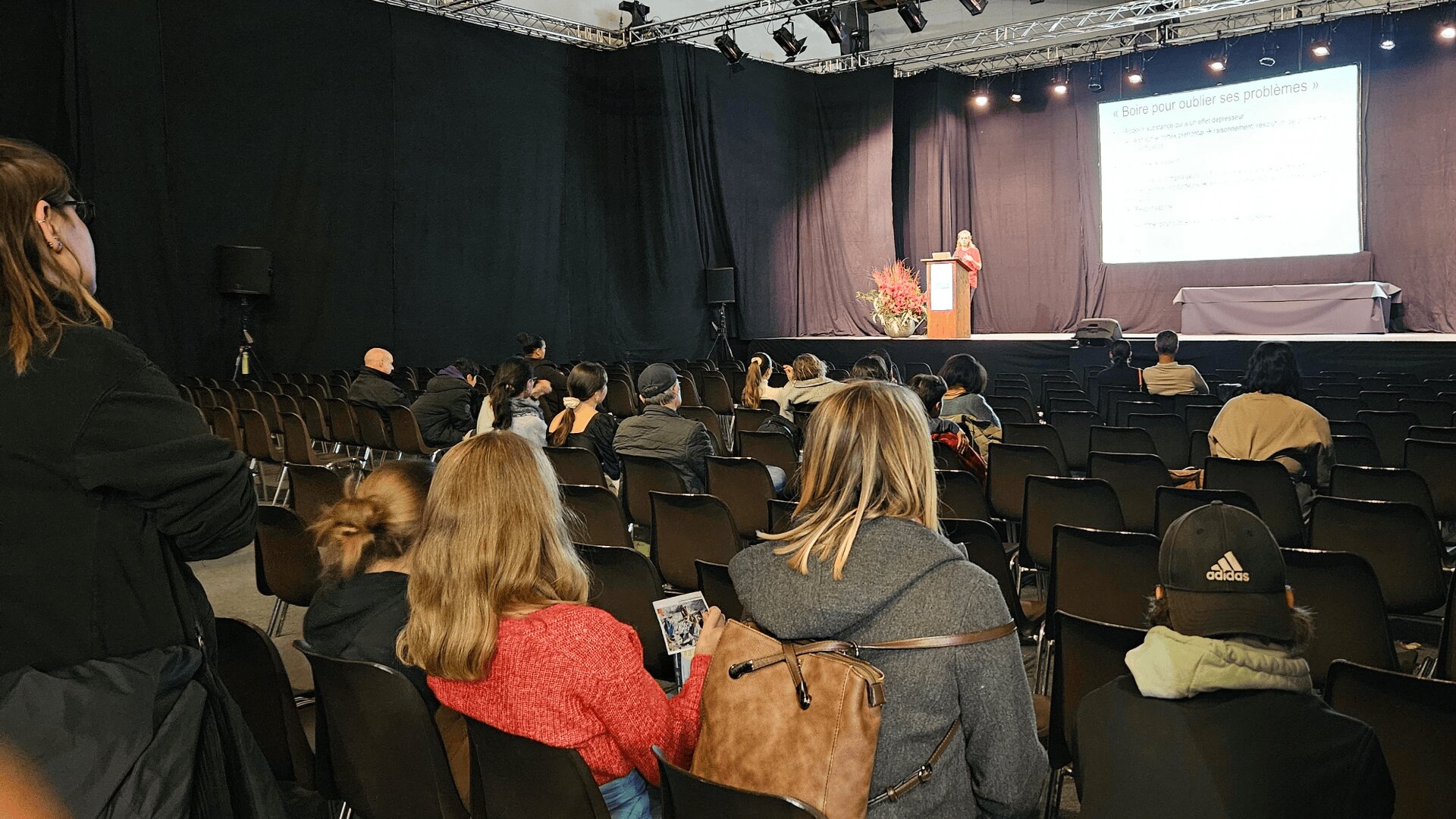 Un orateur se tient sur un podium et s'adresse à un public clairsemé dans une salle de conférence peu éclairée. Un écran de projection affiche un texte en français. Certains membres du public font face à la scène et écoutent attentivement.