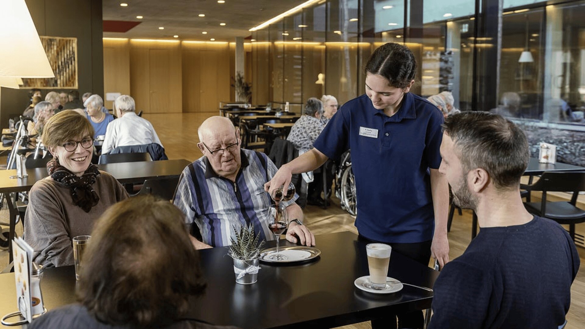 Un serveur verse du vin rouge à un homme âgé assis à une table avec trois autres personnes dans une salle à manger moderne et bien éclairée. D'autres convives sont assis à des tables à l'arrière-plan.