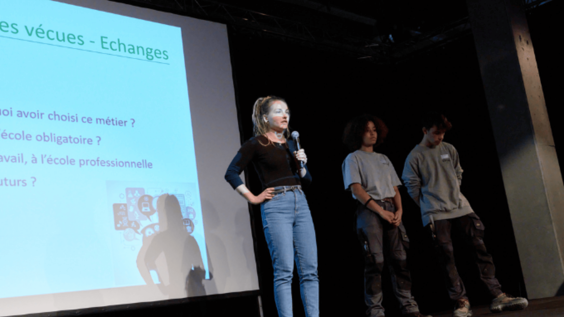 Une femme parle dans un micro sur scène à côté de deux jeunes. Un écran de projection à côté d'eux affiche un texte en français sur les choix de carrière et l'éducation.
