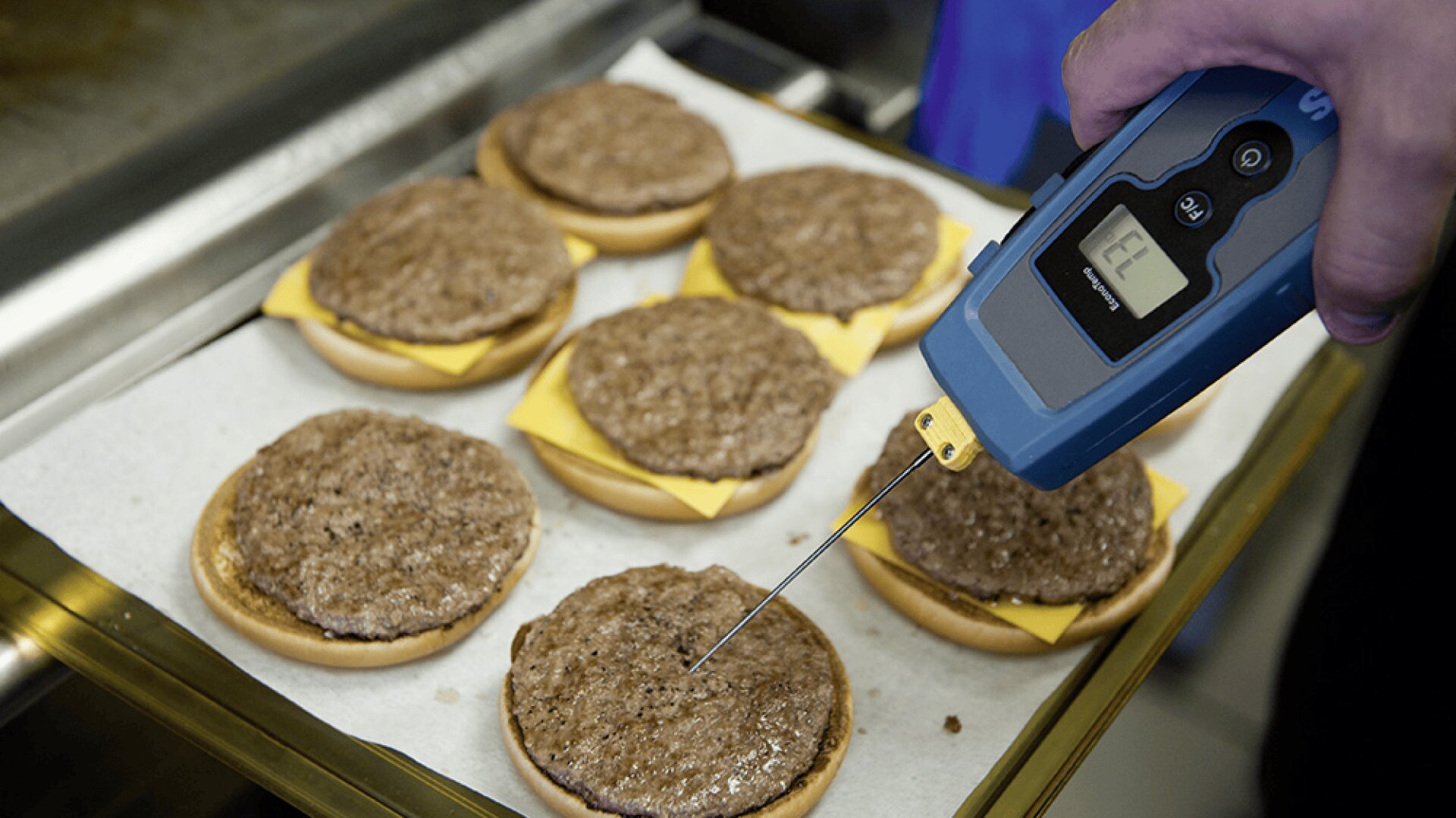 Une personne utilise un thermomètre alimentaire numérique pour vérifier la température des galettes de hamburger cuites placées sur des cheeseburgers ouverts, qui sont disposés sur un plateau tapissé de papier sulfurisé.