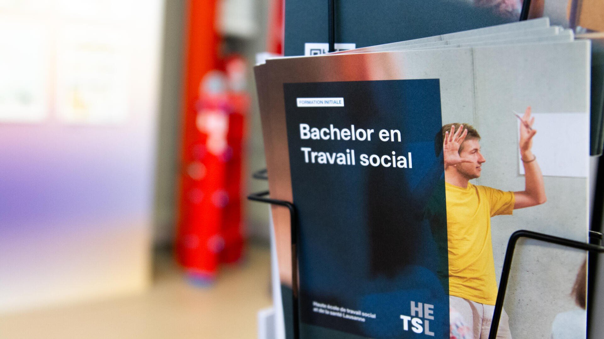 Une brochure intitulée Bachelor en Travail social est présentée dans un présentoir, avec un homme souriant en chemise jaune qui fait des gestes avec les mains levées, sur un fond flou.