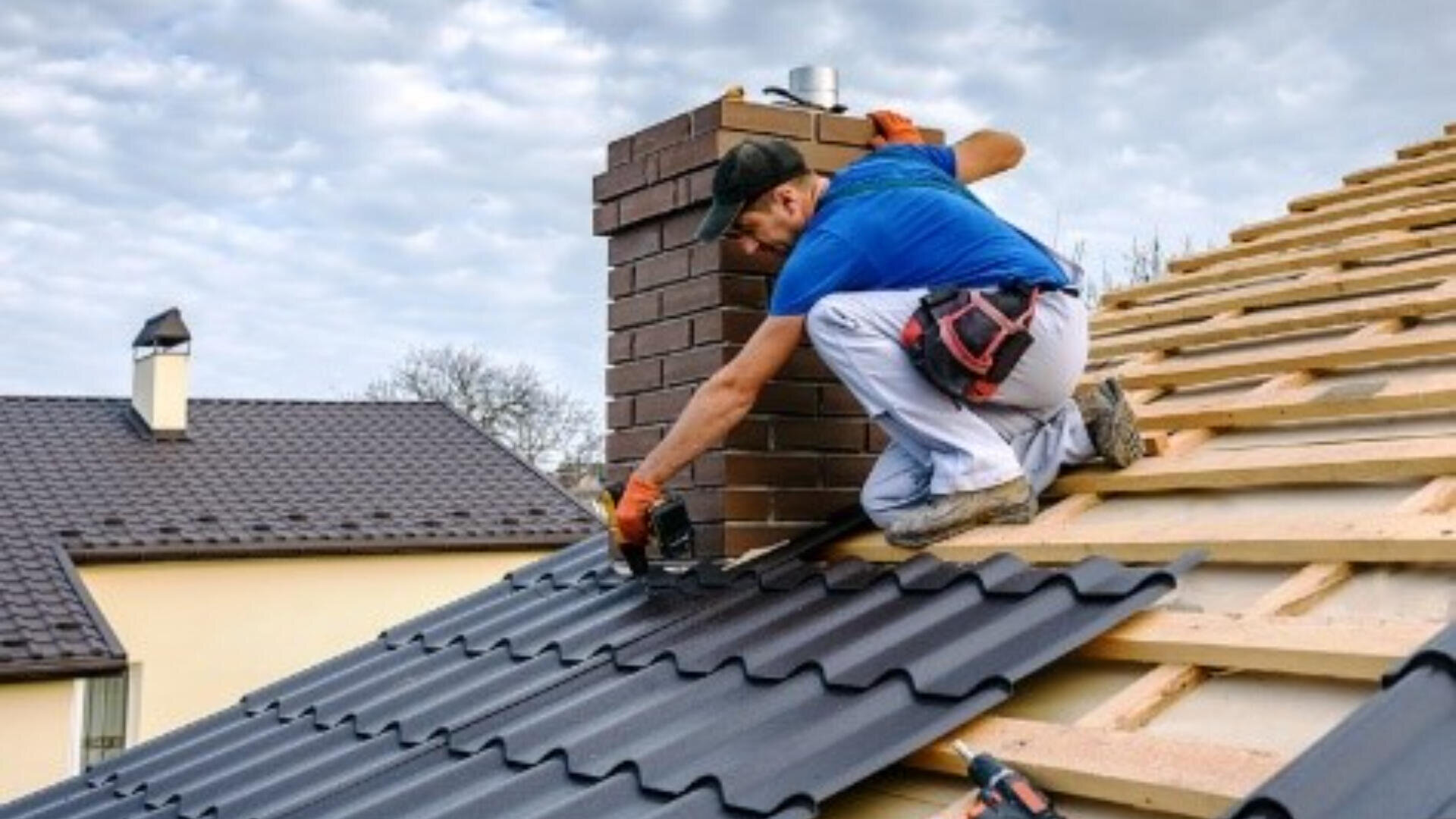 Un ouvrier vêtu d'une chemise bleue et d'une casquette installe des tôles noires sur un toit en bois, à côté d'une cheminée en briques, sous un ciel nuageux. Des maisons et des arbres sont visibles à l'arrière-plan.