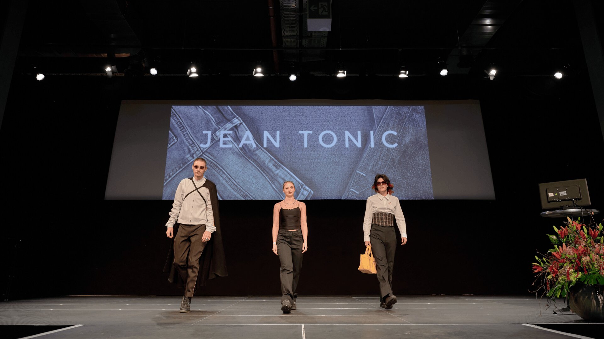 Trois mannequins défilent sur un podium avec JEAN TONIC affiché sur un grand écran derrière eux. Elles portent des vêtements modernes et élégants dans des tons neutres, et des lumières éclairent la scène.