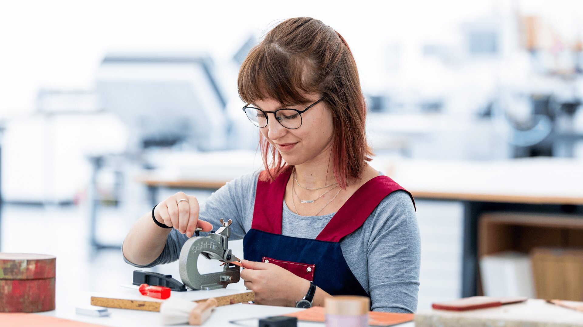 Une femme aux lunettes et aux cheveux bruns travaille avec une petite machine industrielle à une table d'atelier, en se concentrant intensément. Divers outils et matériaux sont éparpillés sur la table autour d'elle.